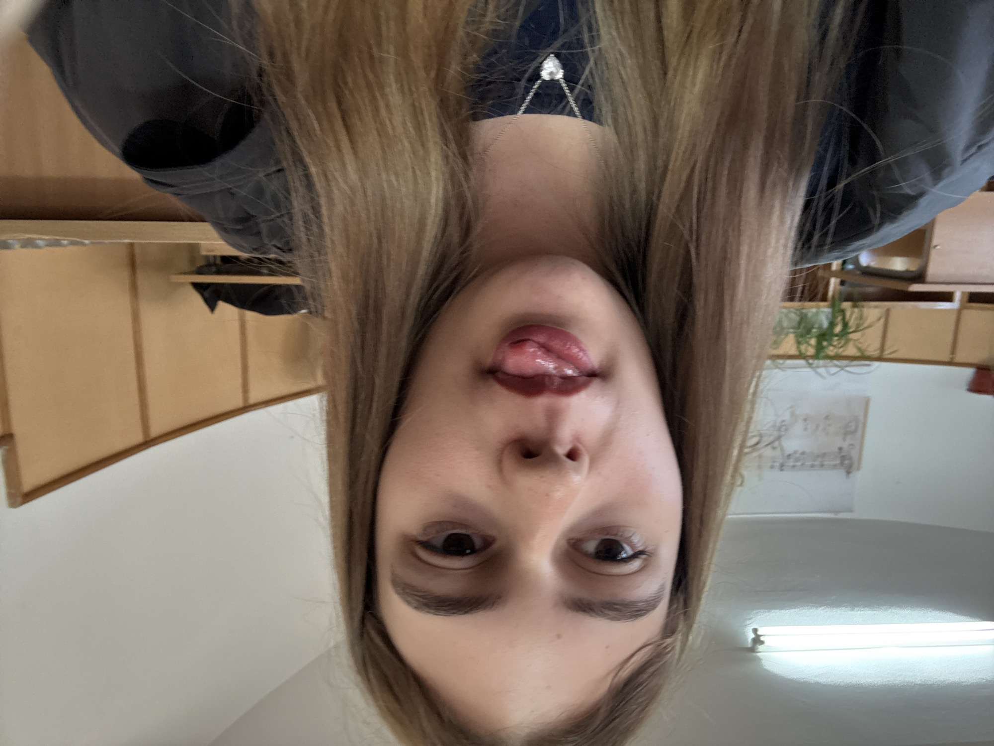 A selfie-style photograph shows a person with long, light brown hair, their face filling the frame. They are sticking their tongue out slightly. The person is wearing a necklace with a pendant. The background shows a room with wooden cabinets, a plant, and a fluorescent light fixture on the ceiling. There is also a piece of paper with writing on it, partially visible. It is not possible to identify the location in Panagyurishte, Bulgaria from the image.