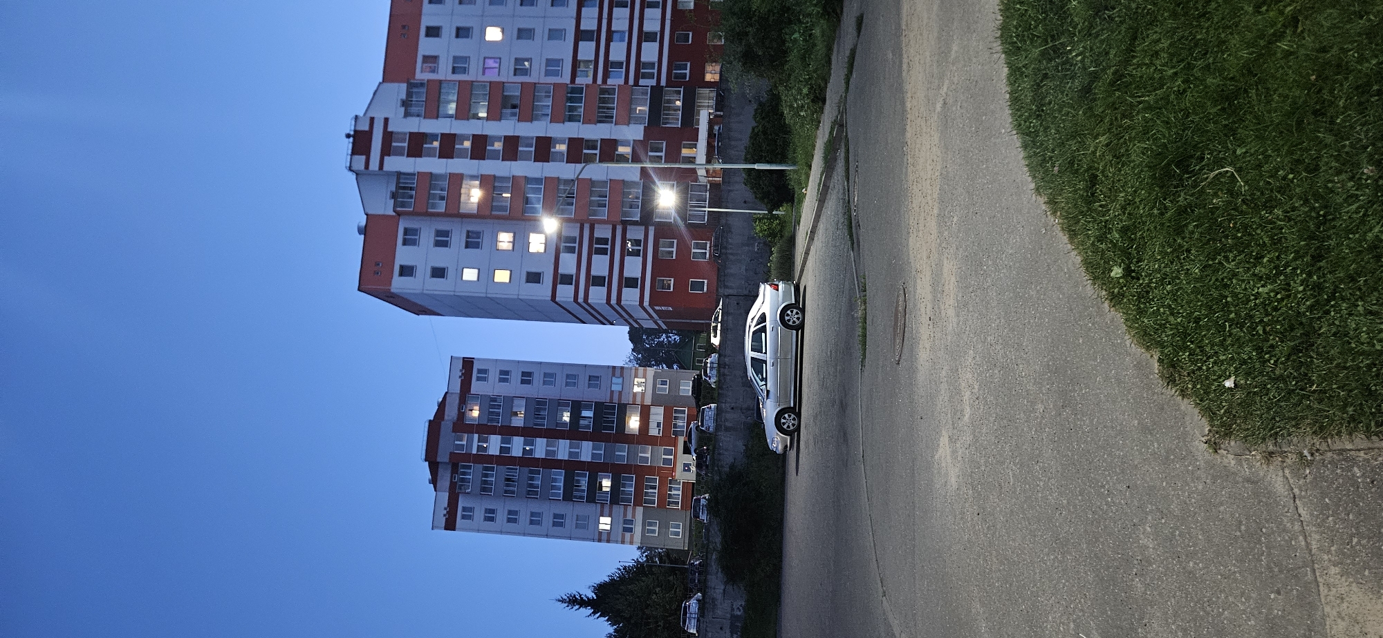 This is a vertical, full-shot, low-angle photograph taken at dusk in Vitsebsk, Belarus. The scene depicts two identical, multi-story apartment buildings with red and white facades, positioned to the left and right sides of the frame, respectively. The building on the left is closer to the viewer. Both buildings have numerous windows, some of which are illuminated. A silver sedan is parked in the foreground on an asphalt lot. The lot slopes downwards towards the right, with a patch of green grass in the lower right corner. A concrete retaining wall runs along the base of the apartment buildings, with a line of parked cars visible behind it. Trees and foliage are present on the left and right sides of the frame, and a street light illuminates the area between the apartment buildings and the parking lot. The sky is a deep blue, indicating twilight. The image appears to be taken from a public space, likely a residential area.