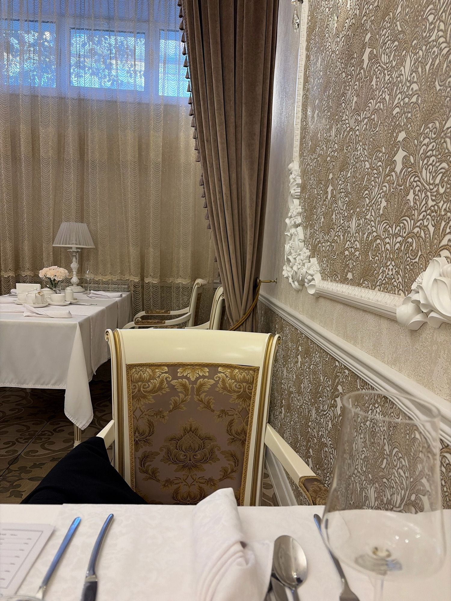 The image depicts an interior scene likely in a restaurant or dining establishment. A formal table setting is visible, complete with white tablecloth, folded napkins, cutlery, and glassware. A white teapot, cups, and a small vase with pink flowers are arranged on the table. The room features ornate wallpaper with a damask pattern in gold and beige tones, complemented by decorative white molding. Plush drapery in a taupe color hangs beside a window with sheer curtains. A lamp with a pleated shade is also present on a side table. The style suggests a sophisticated and classic ambiance. The location is identified as Angarsk, Russia.