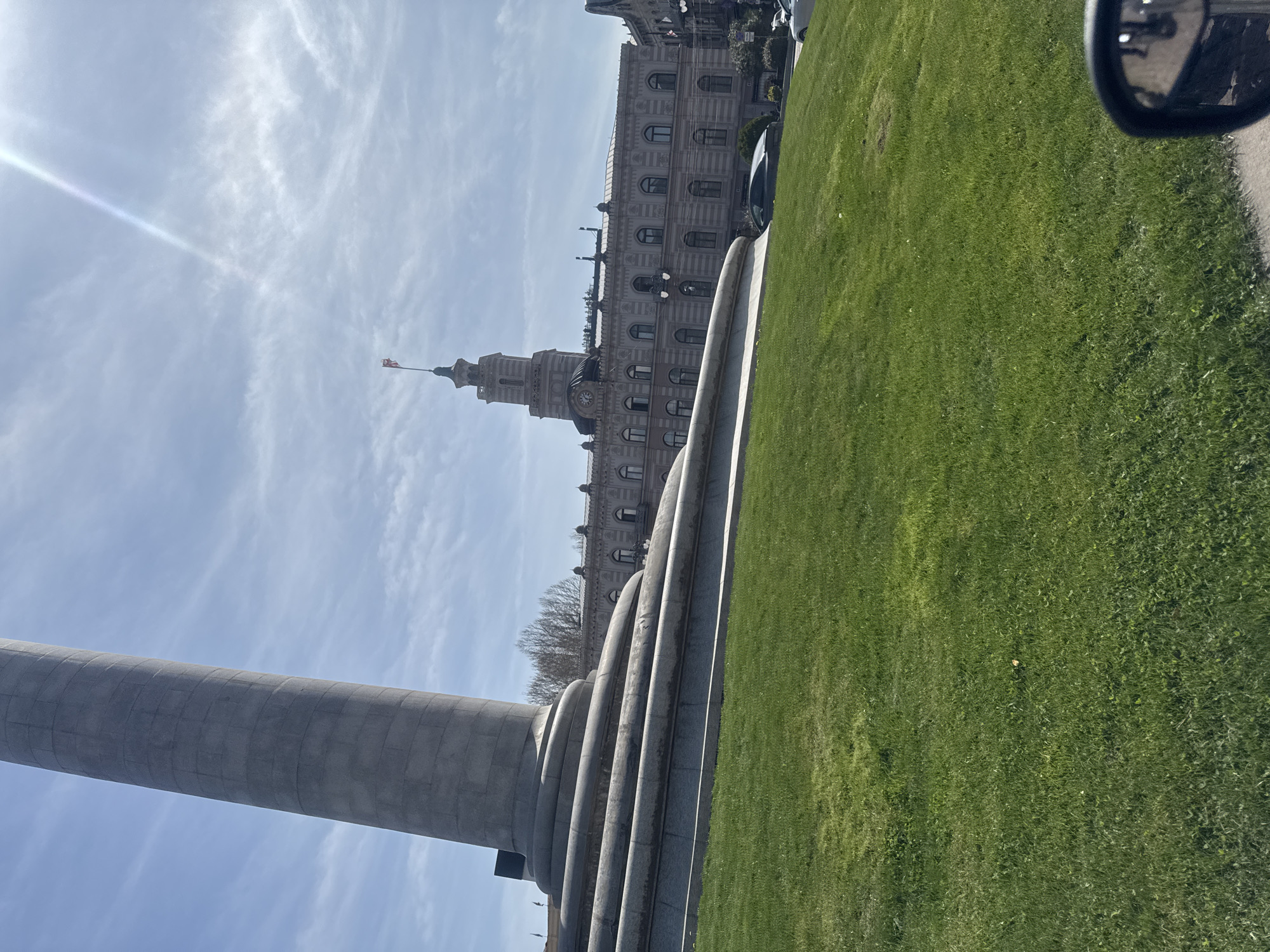 The image shows a view of Freedom Square in Tbilisi, Georgia. A tall gray column dominates the foreground on the left. Behind it is a stone monument with layered curved steps. To the right of the monument stretches a green lawn. In the background, the former City Hall of Tbilisi is visible with its clock tower and flag. The sky is partly cloudy with a visible sun glare. A car side mirror is visible in the top right corner.
