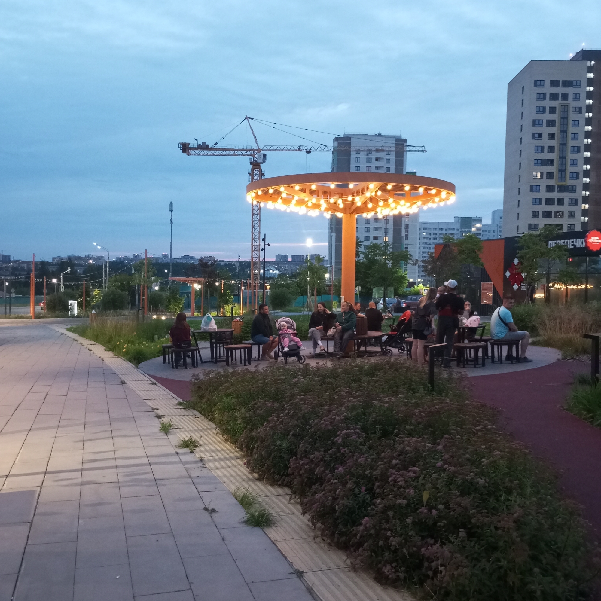 The image captures an outdoor seating area in a park-like setting within the city of Pervomaysky, Russia, during the twilight hours. A circular, open-air canopy with numerous hanging lights illuminates the central seating arrangement. Several individuals are gathered around tables, some seated, others standing. One table has a stroller with a baby. To the left, a paved walkway with textured strips runs alongside a lawn area. In the background, tall residential buildings and a construction crane are visible against a dusky sky. The overall atmosphere suggests a casual gathering or dining experience.