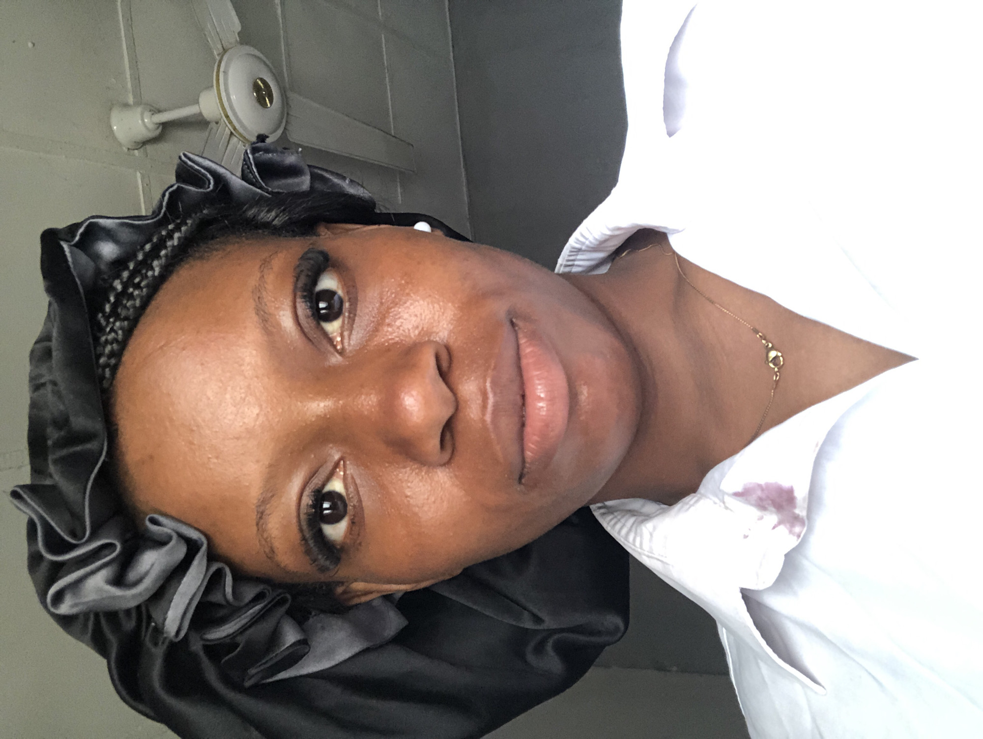 A medium shot portrait shows a Black person wearing a black satin bonnet and a white collared shirt. The person's face is angled slightly to the right, and they are looking directly at the camera. Their hair is braided, and the bonnet covers the sides and back of their head. The white shirt has a small, dark stain on the collar. A gold necklace with a small, heart-shaped pendant is visible around their neck. In the background, a white ceiling fan is attached to a white ceiling. The lighting suggests an indoor setting, likely a home. The image does not provide specific information about the location within Kashere, Nigeria, but it depicts a common domestic scene.