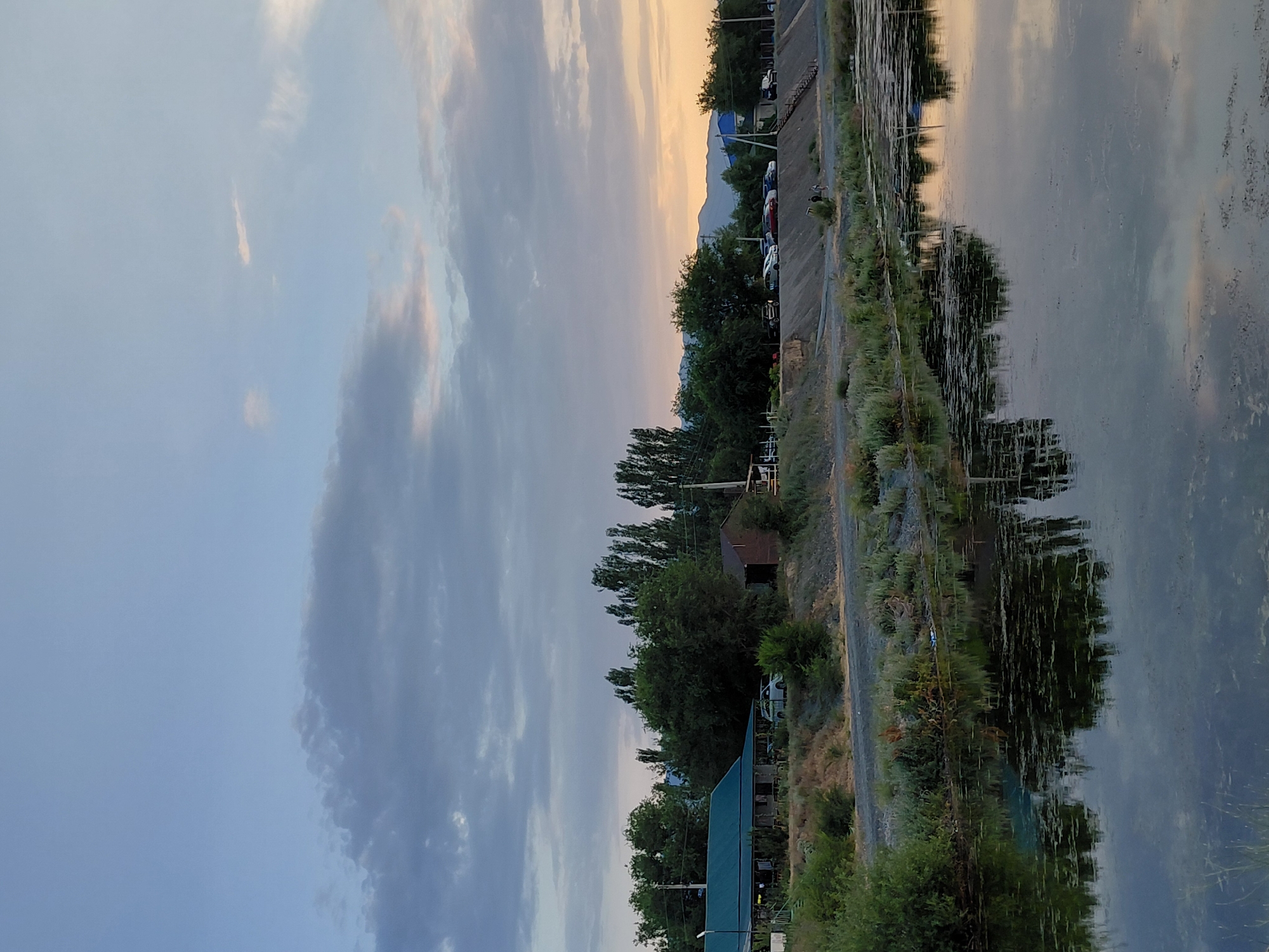 The image depicts a waterscape at dusk with a view towards a landmass. A calm body of water, likely a lake or river, dominates the right side of the frame, reflecting the sky and surrounding vegetation. The water's surface is textured with darker patches, possibly indicating aquatic plants or debris.
To the left of the water, a sloped embankment is visible, covered with sparse vegetation and a gravel path. Further inland, a line of trees, mostly deciduous, forms a natural border. Among the trees, several structures are partially visible: a small, rustic shed with a wooden exterior and a larger building with a green, sloped roof, suggesting a park or recreational area.
Along the path and slightly elevated, a row of parked vehicles, including cars and possibly a truck, can be seen. The background reveals a mountainous landscape, its silhouette softened by the twilight. The sky is a canvas of dramatic clouds, with hues of pale blue, gray, and hints of warm orange and pink from the setting sun.
The time of day is clearly evening, as indicated by the soft light and the sky's colors. The weather appears calm, with no signs of wind or precipitation. The scene evokes a sense of tranquility and natural beauty. No people are explicitly visible in the image. The location context is Ақши, Kazakhstan, which could suggest a rural or semi-rural setting with natural water features. No text is visible in the image.