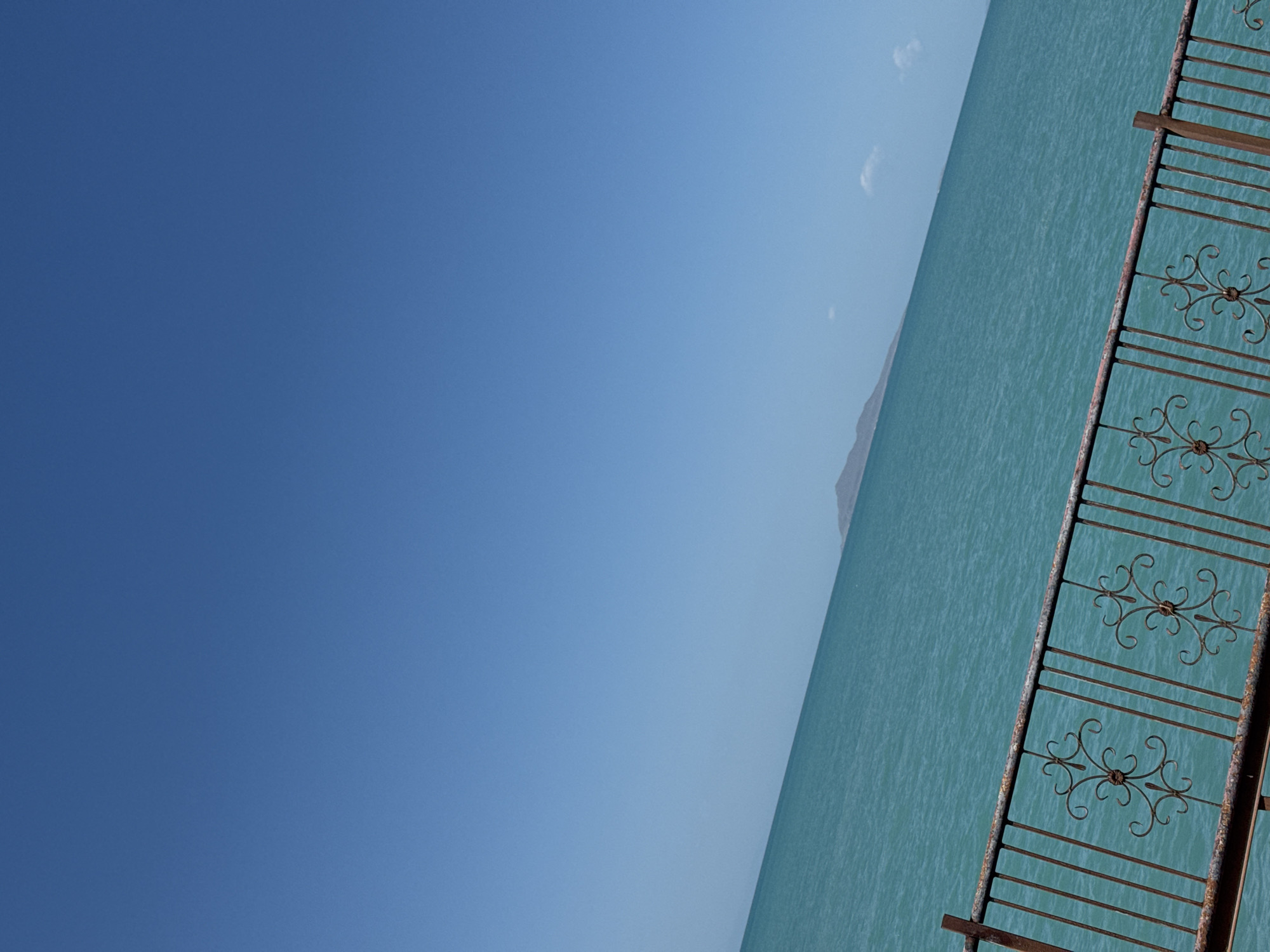 The image displays a wide expanse of turquoise water under a clear blue sky. In the foreground, a rusty metal railing with ornate scrollwork runs diagonally across the frame, suggesting a waterfront vantage point. The railing is weathered and shows signs of age. The water stretches towards a distant, low-lying island or landmass with a muted, brownish-gray hue. The horizon is not clearly defined due to the angle of the shot and the vastness of the water. The scene is likely captured on a sunny day, given the brightness and the clear sky. The location is identified as Zharbulak, Kazakhstan, implying this is a view of a body of water in or near that region. There are no people or specific actions depicted in the image.