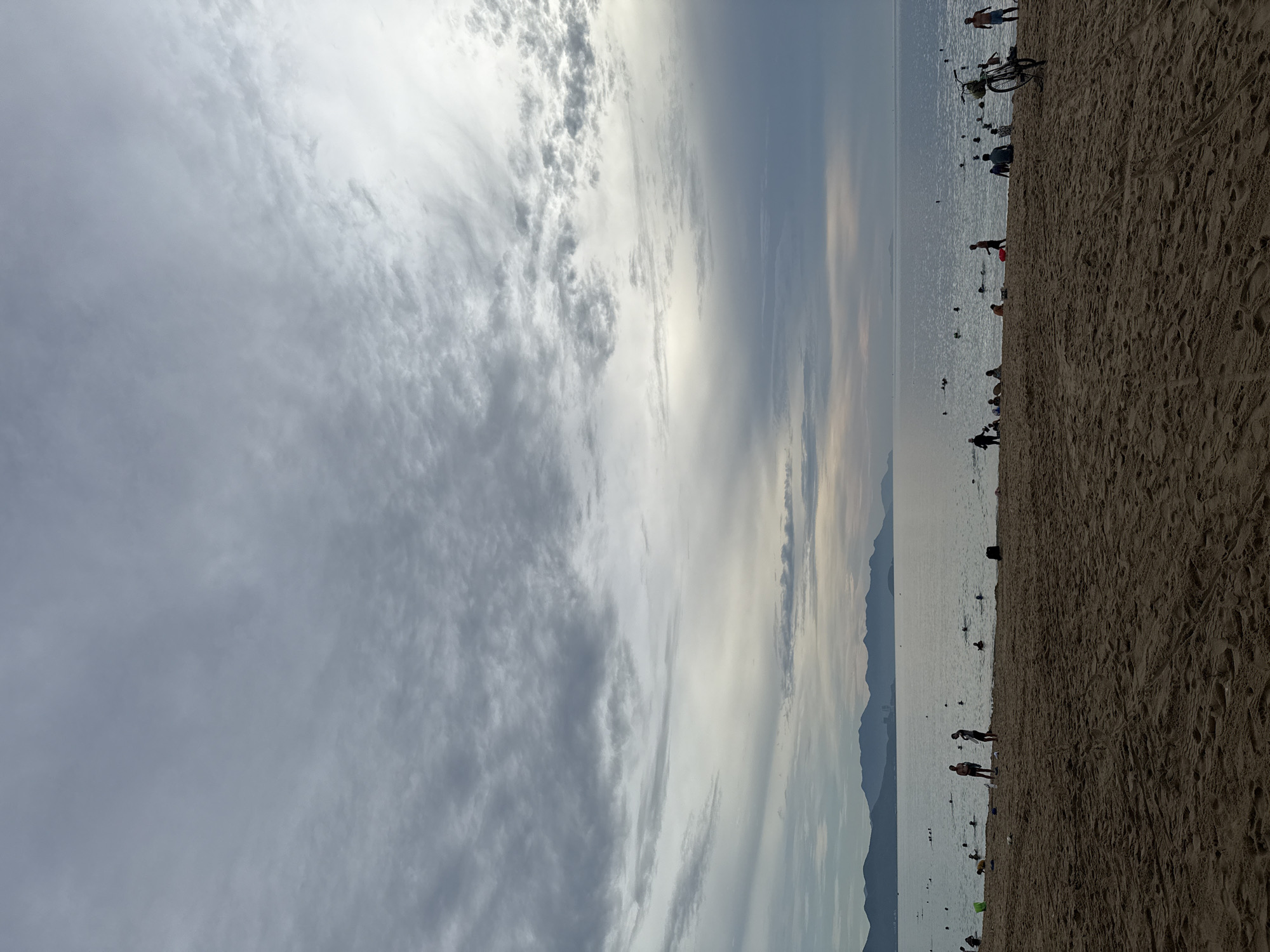 This image captures a beach scene in Nha Trang, Vietnam, during what appears to be late afternoon or early evening due to the overcast sky and subdued lighting. The foreground is dominated by sandy beach with footprints and tire tracks visible. A cluster of people are scattered across the sand, some standing, some sitting, and a few are walking towards the water.
In the middle ground, the ocean stretches out, dotted with numerous people swimming and wading in the shallow water. The water is calm with gentle ripples.
In the background, a silhouette of mountains forms the horizon line. The sky is filled with dramatic, heavy gray clouds, with lighter patches indicating a break in the cloud cover or the fading sunlight. A bicycle is visible on the right side of the beach, leaning near the water's edge. The overall atmosphere is one of a relaxed beach day, with people enjoying the water and the shore under a cloudy sky.