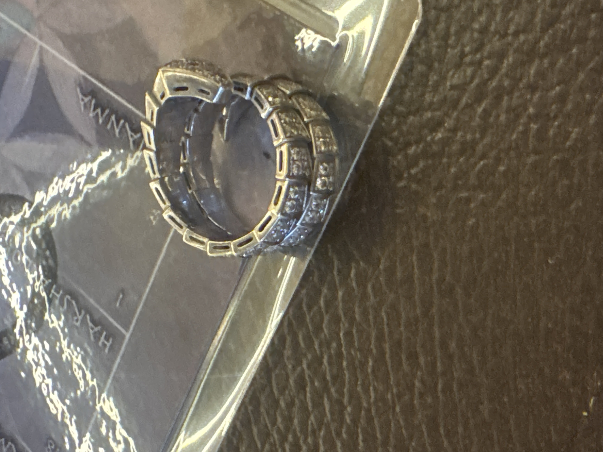 The image shows a silver-colored ring with small, square-cut stones set into it. The ring appears to be resting on a transparent plastic package, upon which are partially visible words like "ANNA" and "HARSH." The background is a textured brown surface. The location is North Kuta, Indonesia.