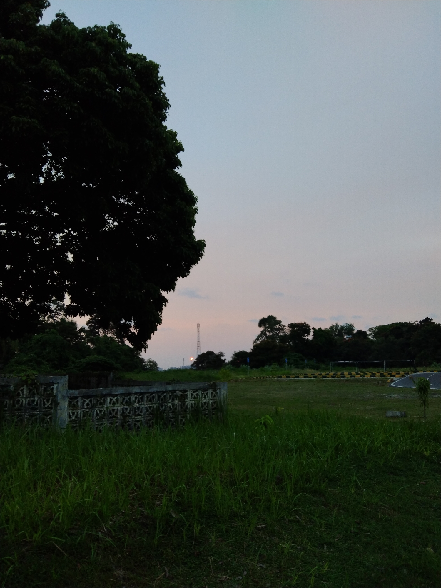 The image depicts an outdoor scene in Lhokseumawe, Indonesia, captured during either dusk or dawn. The sky presents a soft, ethereal gradient, transitioning from a light blue overhead to a warm, faint pink and orange hue near the horizon. This muted lighting casts a tranquil ambiance over the landscape.
A large, dense tree dominates the left side of the frame, its dark foliage silhouetted against the subtly lit sky. In the immediate foreground, a patch of vibrant green grass grows tall. Behind this, a low, weathered concrete wall or fence stretches horizontally across the middle left, featuring decorative, repeating patterns of cutouts.
Beyond the wall and extending into the mid-ground is an open, grassy field. To the right of the field, a paved track or road is visible, lined with distinctive yellow and black barriers, suggesting a recreational facility or a driving practice area. In the far distance, slightly to the right of the center, a tall, slender communication tower rises against the horizon, with a small light illuminated near its base, further indicating the low light conditions. A line of smaller trees and bushes forms a dark treeline in the mid-ground.
No people or explicit activities are visible, contributing to the serene and still nature of the scene. The overall impression is one of peaceful twilight in a semi-urban or rural setting. There is no visible text in the image.
