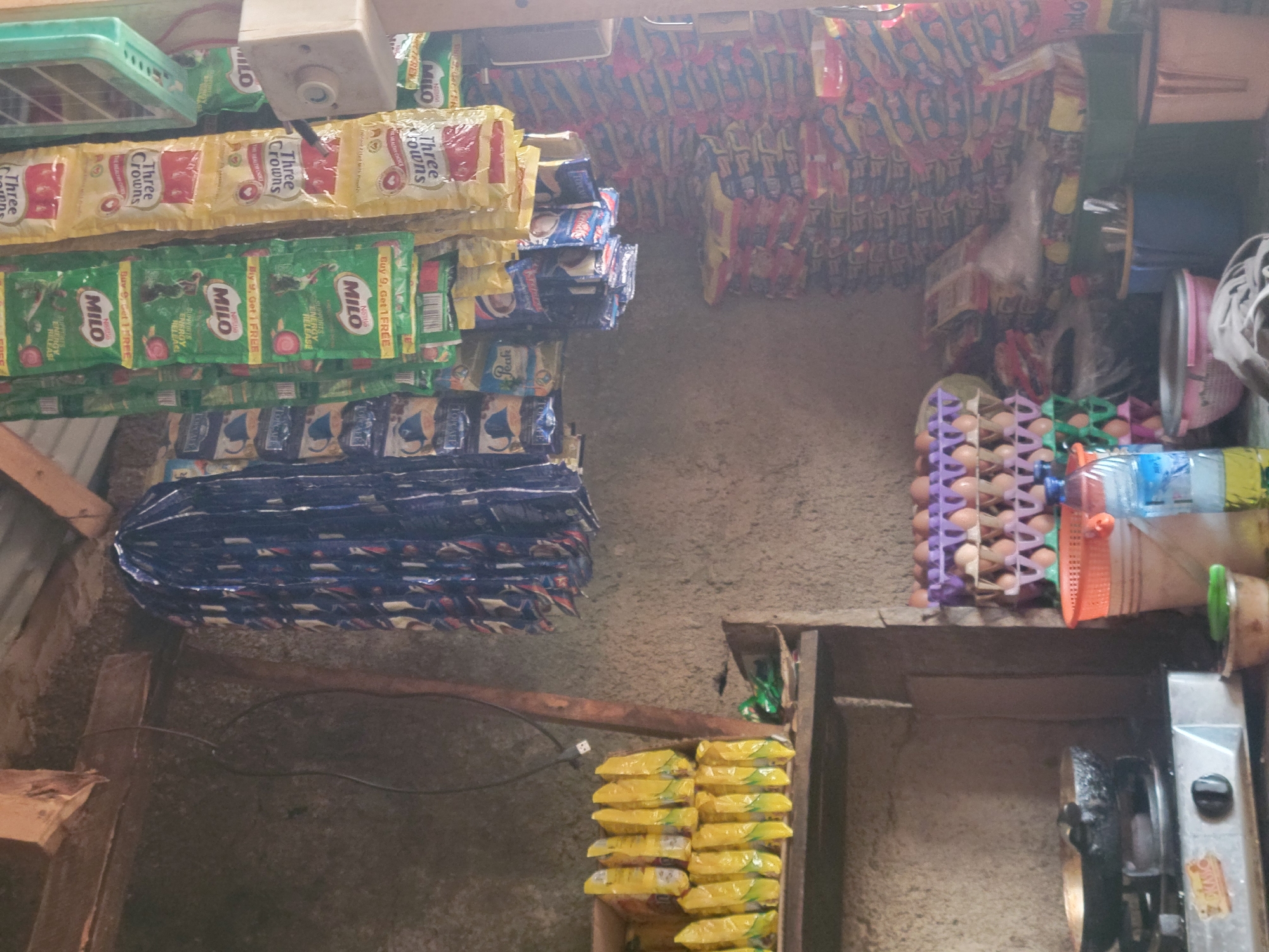 The image depicts the interior of a small convenience store in Madalla, Nigeria. The store appears to be cluttered, with shelves and hanging displays stocked with various food items and beverages. In the foreground, a gas stove with a pan on it suggests food preparation. To the right, stacks of packaged goods line the walls, some of which are identifiable as Milo and Three Crowns products, common brands in Nigeria. An egg carton filled with eggs is also visible, along with buckets and bottles of water. The store's walls are unplastered, revealing a rough concrete or mud texture. The ceiling is supported by wooden beams. The overall impression is of a modestly stocked local shop. There are no people visible in the image.