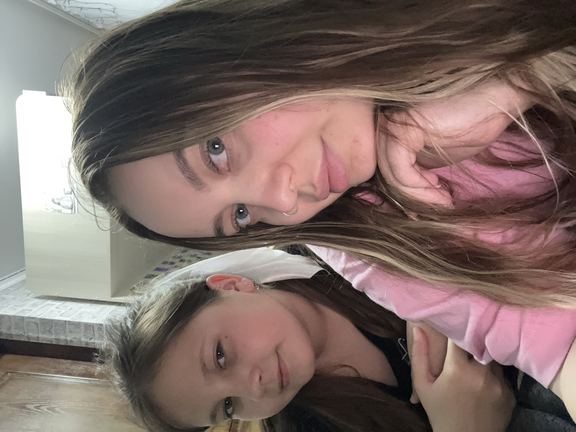 Two females, one a young girl and the other a young adult, pose for a selfie-style photograph indoors. The young adult, positioned higher in the frame, has long brown hair with lighter highlights, light-colored eyes, and a nose ring. She wears a pink garment and appears to be touching her face with one hand. Below her, the young girl has long brown hair and dark eyes. The background includes a cream-colored cabinet or structure and a tiled surface, possibly a countertop or backsplash. The location is Novopokrovskaya, Russia.