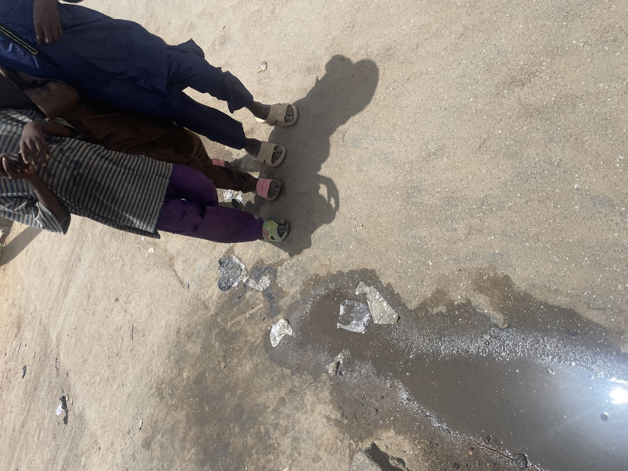 The image shows a street scene in an urban environment, possibly in Unknown, Unknown. Three children are standing at the edge of a puddle or a wet patch on the paved road. The children are wearing casual clothes and sandals. The road surface is rough and appears to be concrete or asphalt. Scattered debris and litter are visible around the puddle. The sunlight is bright and creates sharp shadows.