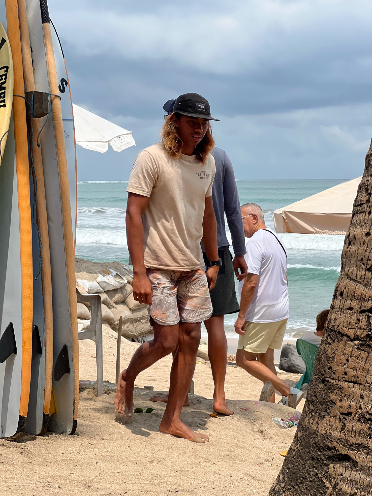 The image depicts a beach scene in Kuta, Indonesia, likely a popular surf spot. In the foreground, several surfboards are leaning against a structure. A young man with long, blonde hair and a black baseball cap is walking towards the right, wearing a tan t-shirt with text on it and patterned shorts. Beside him, another person in a blue long-sleeved shirt and dark shorts is also walking. Further to the right, an older man in a white t-shirt and light yellow shorts is walking away from the camera, with a black strap across his shoulder. In the background, the ocean with waves is visible, along with beach umbrellas and sandbags. The sand is light brown, and the sky is cloudy. The overall atmosphere suggests a casual day at the beach, possibly related to surfing activities.