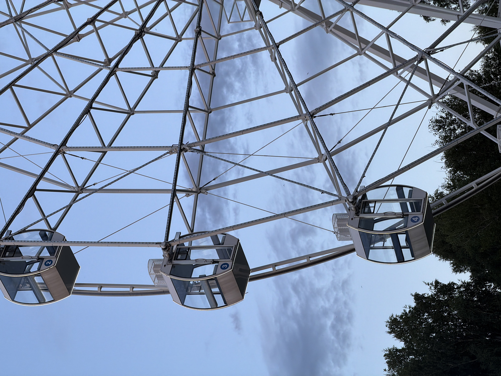 This is a low-angle, rotated photograph of a Ferris wheel in Nizhnekamsk, Russia. The subjects are the gondolas of the Ferris wheel, which are enclosed, hexagonal structures with glass windows and metal framing. There are at least four gondolas visible in the image, numbered 2, 3, and 4. The Ferris wheel's metallic structure is intricate, with a lattice of beams and supports against a cloudy sky. Trees are visible on the right side of the image, suggesting an outdoor setting, possibly a park or amusement area. The lighting suggests it is either dawn or dusk, with a soft, diffused light. The sky is a pale blue with streaks of grey clouds, indicating overcast or partly cloudy weather. The overall mood is calm and serene, with the Ferris wheel appearing stationary. There are no people visible in the gondolas. The image captures the scale and geometric complexity of the Ferris wheel's construction.