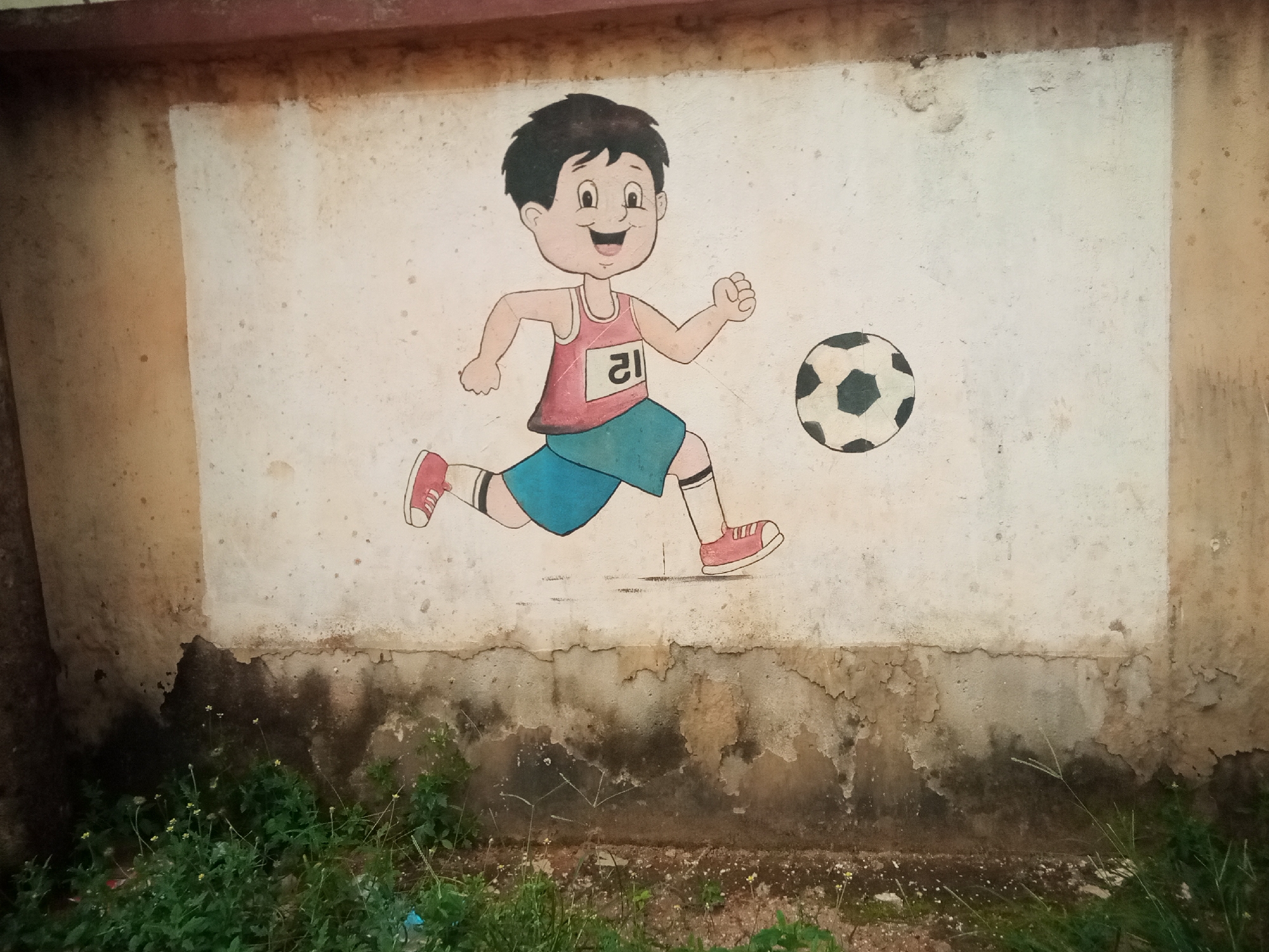 In Oko Town, Nigeria, a faded mural painted on a weathered concrete wall depicts a cartoon boy in mid-stride, kicking a soccer ball. The boy wears a red tank top with the number "21" in a white box, blue shorts, and red sneakers. He has black hair and a cheerful expression. The soccer ball is black and white, with hexagonal and pentagonal panels. The wall itself shows signs of age and wear, with peeling paint and water stains, particularly at the lower portion where it meets a patch of green foliage and sparse wildflowers. The overall impression is of a once-vibrant artwork now showing the effects of time and exposure to the elements in an outdoor setting.