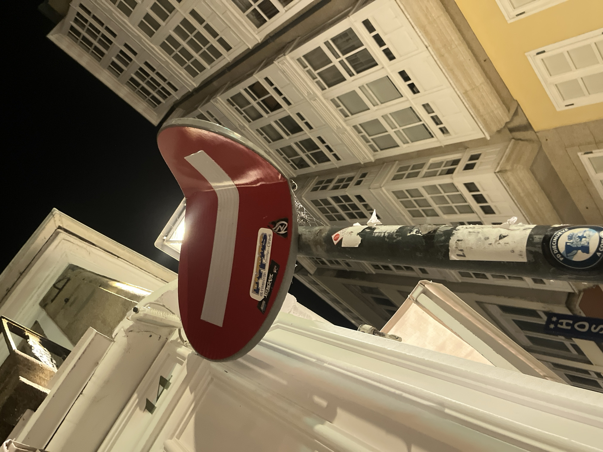 The image, captured at night, presents a low-angle view of a no-entry traffic sign mounted on a dark, cylindrical pole. The sign is red with a horizontal white bar. Several stickers adorn the sign, including one with text that appears to read "LAVIPAS" and another with "TORCAS." The pole itself is weathered and covered in various papers and stickers, one circular sticker at the bottom has a blue border with an emblem and text that seems to read "PD DIECINUEVE A COMU."
Behind the sign, a white building with numerous windows is visible, characteristic of the architecture found in A Coruña, Spain. The windows are framed in white and set into the building's facade, which is also predominantly white. The building's design, with its enclosed balconies or "galerías," is a distinctive feature of this Galician city. To the right of this building, another structure with a bright yellow facade is partially visible, also featuring white-framed windows.
The overall setting suggests an urban environment, likely a street in A Coruña, given the architectural style. The lighting indicates it is nighttime, with artificial lights illuminating the buildings and the street sign. The perspective is tilted upwards, making the buildings appear to loom over the viewer.