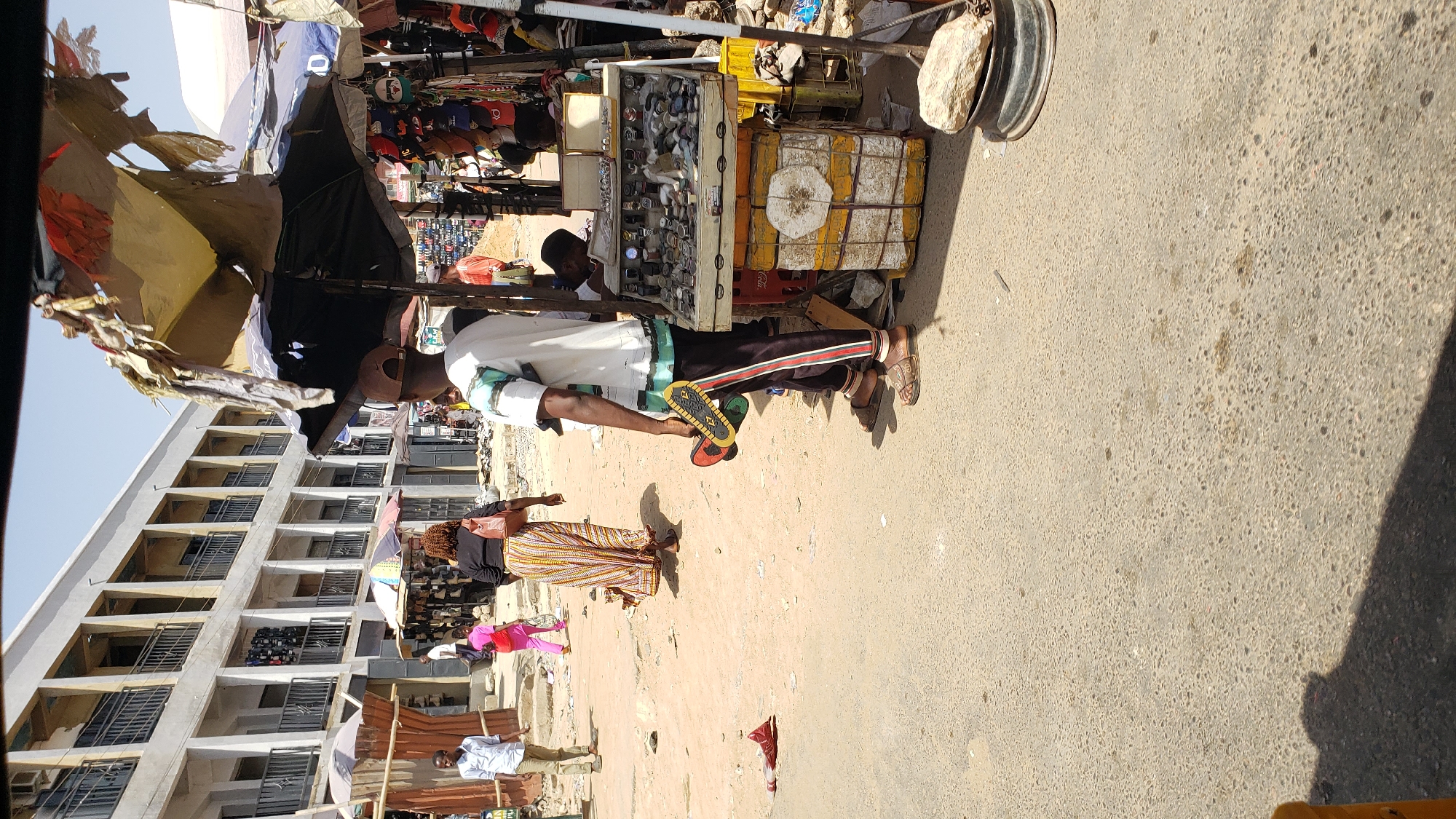 The image depicts an outdoor street scene in Maiburiji, Nigeria, with a man standing next to a makeshift shop. The man is wearing a white shirt, black pants with stripes, and sandals. He is holding a pair of sandals in his hand. The shop is a collection of stacked containers and boxes, showcasing items like watches. Behind the man and the shop, there are other people walking on a sandy ground, and a building under construction is visible in the background. The ground is paved with light-colored concrete.