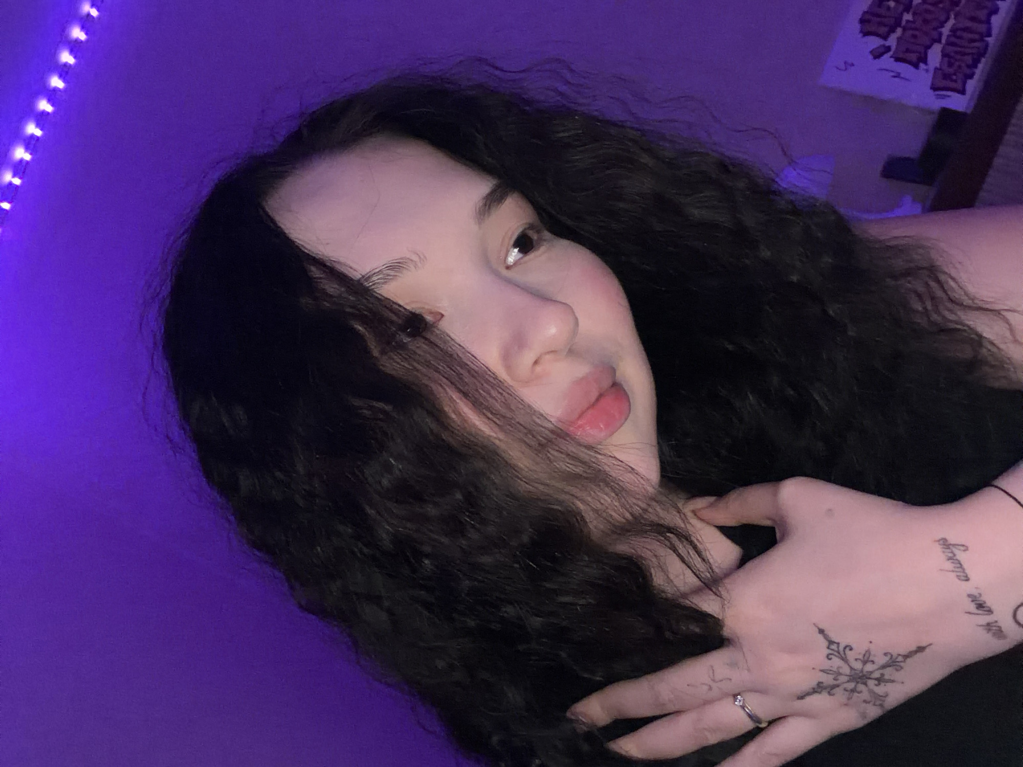 The image features a young individual, likely female, in a close-up shot under purple ambient lighting. The subject has long, dark, wavy hair that cascades around the face and shoulders. The head is tilted downwards and to the right, with the visible right eye looking towards the bottom right. The lips are slightly parted and have a pinkish-red color. The left hand is raised, with fingers gently touching the neck/chin area. On the fourth finger of the left hand, there is a silver-toned ring with a small, clear stone. The back of the left hand features a tattoo of a complex, symmetrical design resembling a stylized snowflake or starburst. Further down on the wrist, another tattoo displays the text "with love, always" in script. The background consists primarily of a solid purple wall. In the upper right corner, a strip of individual purple LED lights is visible, indicating the source of the room's illumination. In the lower right quadrant, a dark, horizontal surface, possibly a shelf or desk, holds a white object with dark, illegible graffiti-style lettering, likely a poster or piece of art. The scene provides no visual cues to suggest its location is in the city of Chaikovsky, Russia.