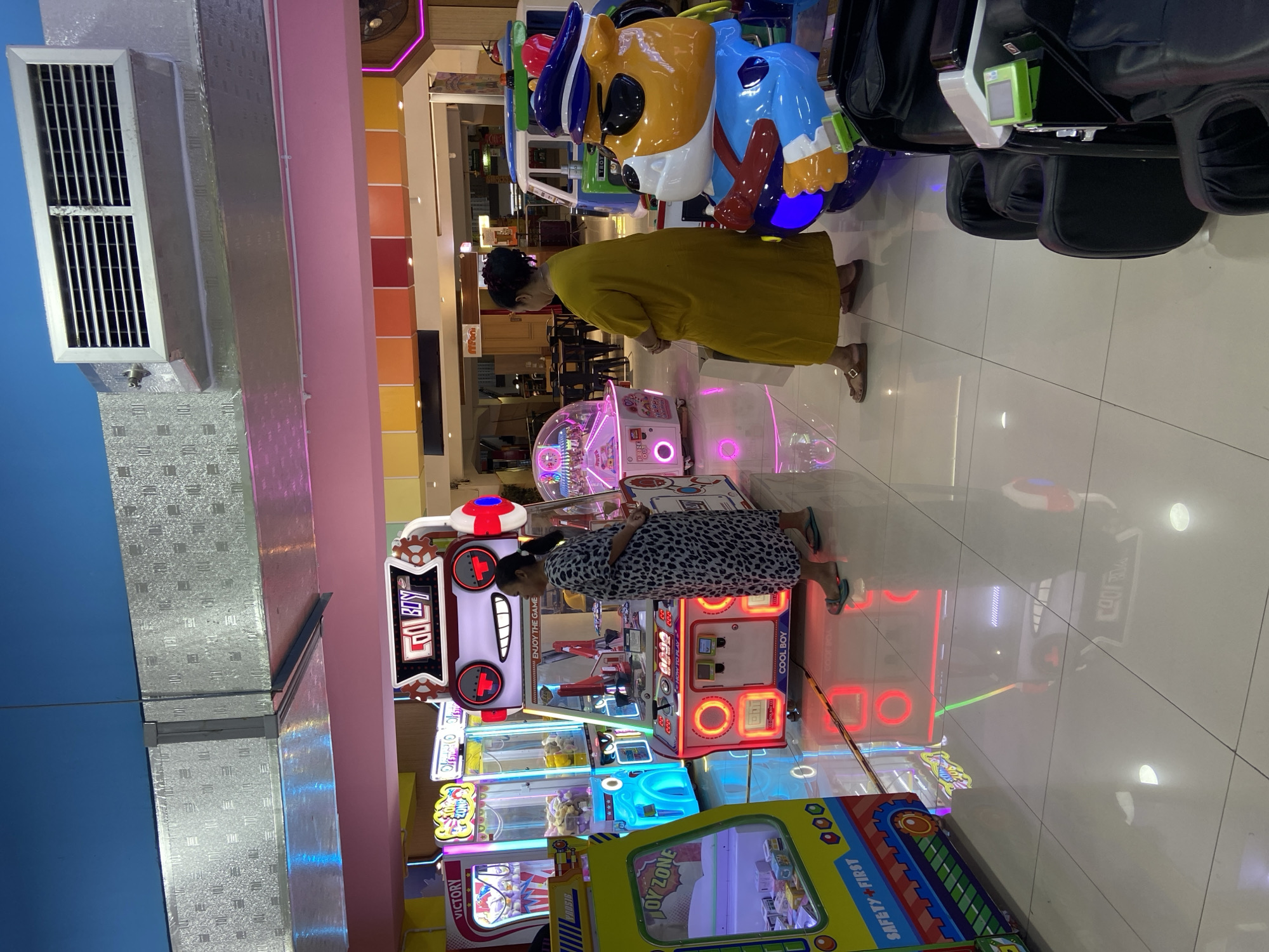 The image depicts an indoor arcade in Kecamatan Pati, Indonesia, featuring rows of colorful arcade machines and a few patrons. In the foreground, a large yellow claw machine with "TOY ZONE" and "SAFETY FIRST" branding is visible on the left. To the right of the claw machine is a "COOL BOY" arcade game, a grabber-style machine with a cartoon robot face, where a woman in a patterned dress is standing. Further back, other arcade machines like "VICTORY" and a pink "KIMPY" machine are present. Another woman in a yellow dress is standing to the right of the "COOL BOY" machine, next to a large, cartoonish rideable toy resembling a police bear in a blue uniform. The arcade has a tiled floor that reflects the bright lights of the machines. In the background, there are tables and chairs, suggesting a seating area. The overall environment is brightly lit with neon and LED lighting, common in entertainment venues. The ceiling features exposed metallic ductwork and colorful ceiling panels.