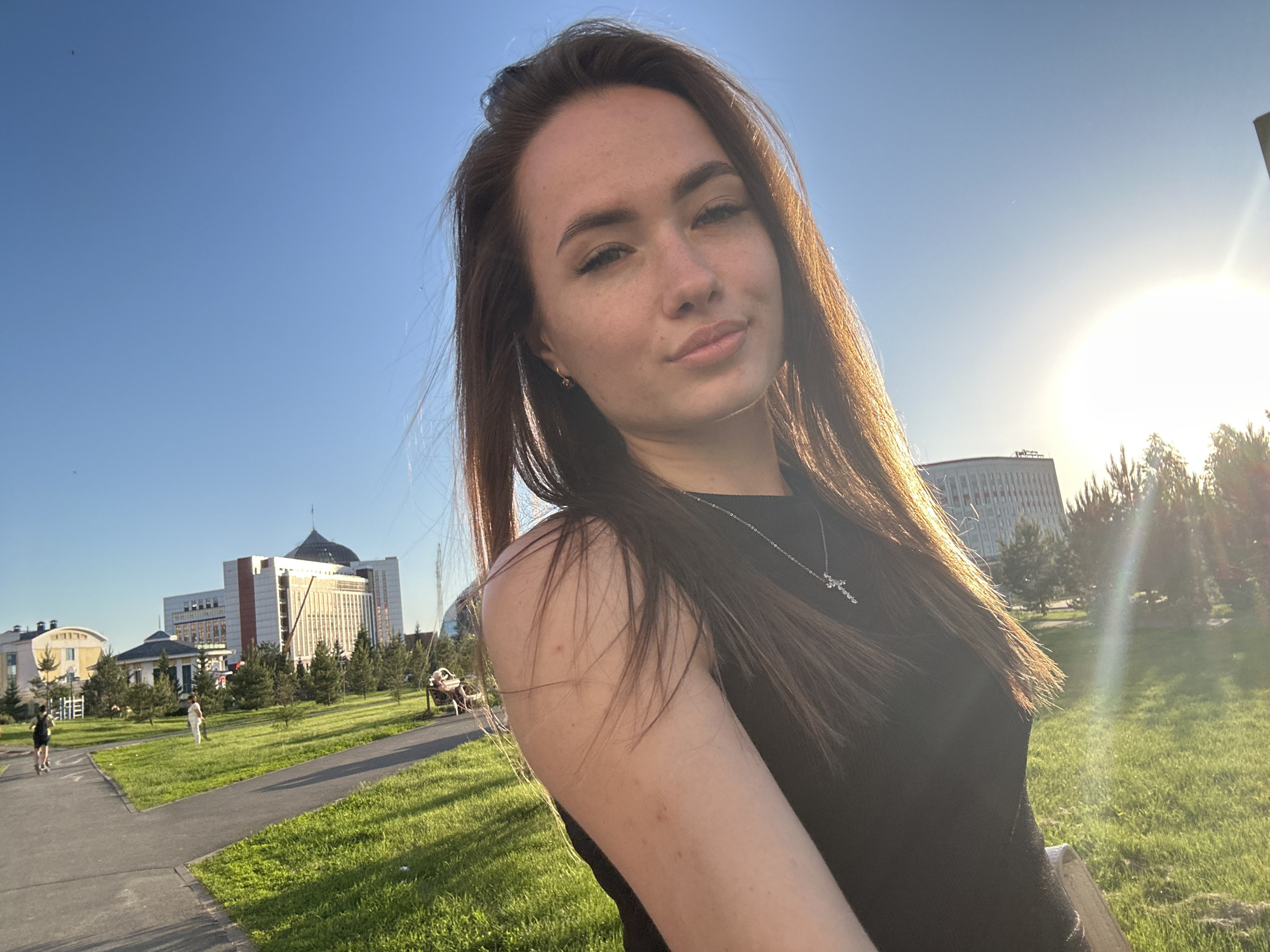The media file presents an outdoor portrait of a young woman. She has long, dark brown hair, fair skin with visible freckles, and a subtle, natural smile directed towards the camera. She is wearing a dark, sleeveless top and a silver necklace adorned with a cross pendant. Her hair is illuminated by the sun, creating golden highlights.
The scene is set in an urban park or green space under a clear blue sky. The time of day appears to be late afternoon or early evening, characterized by the warm, golden light and a strong sun flare emanating from the upper right, indicating the sun's low position. The background features well-maintained green grass, paved walking paths, and various trees, including some conifer-like species.
In the mid-ground and background, several modern buildings are visible, suggesting an urban environment. One prominent building has a distinctive dome or rounded roof, while another displays a contemporary design with large windows. On one of the paths in the distance, two other people can be seen: one individual on a scooter and another walking. No visible text is present in the image.