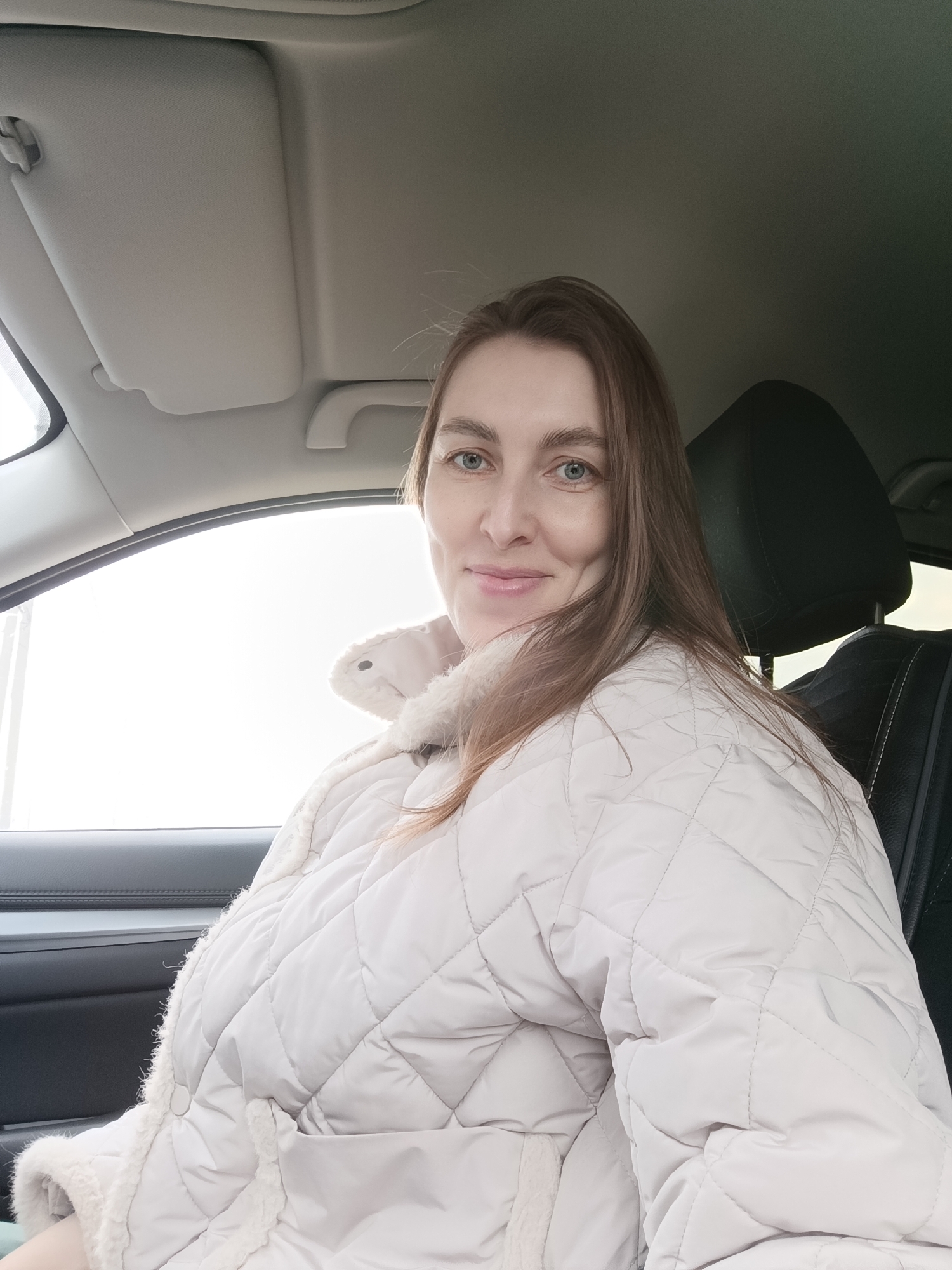 The image depicts a woman in a car. She is wearing a light-colored, quilted jacket with a fur-like trim around the collar. She has light brown hair and blue eyes and is smiling slightly. The interior of the car is visible, including the dashboard, headrest, and window. The outside of the window is bright and blurry. The image was taken in the city of Chaikovsky, Russia.