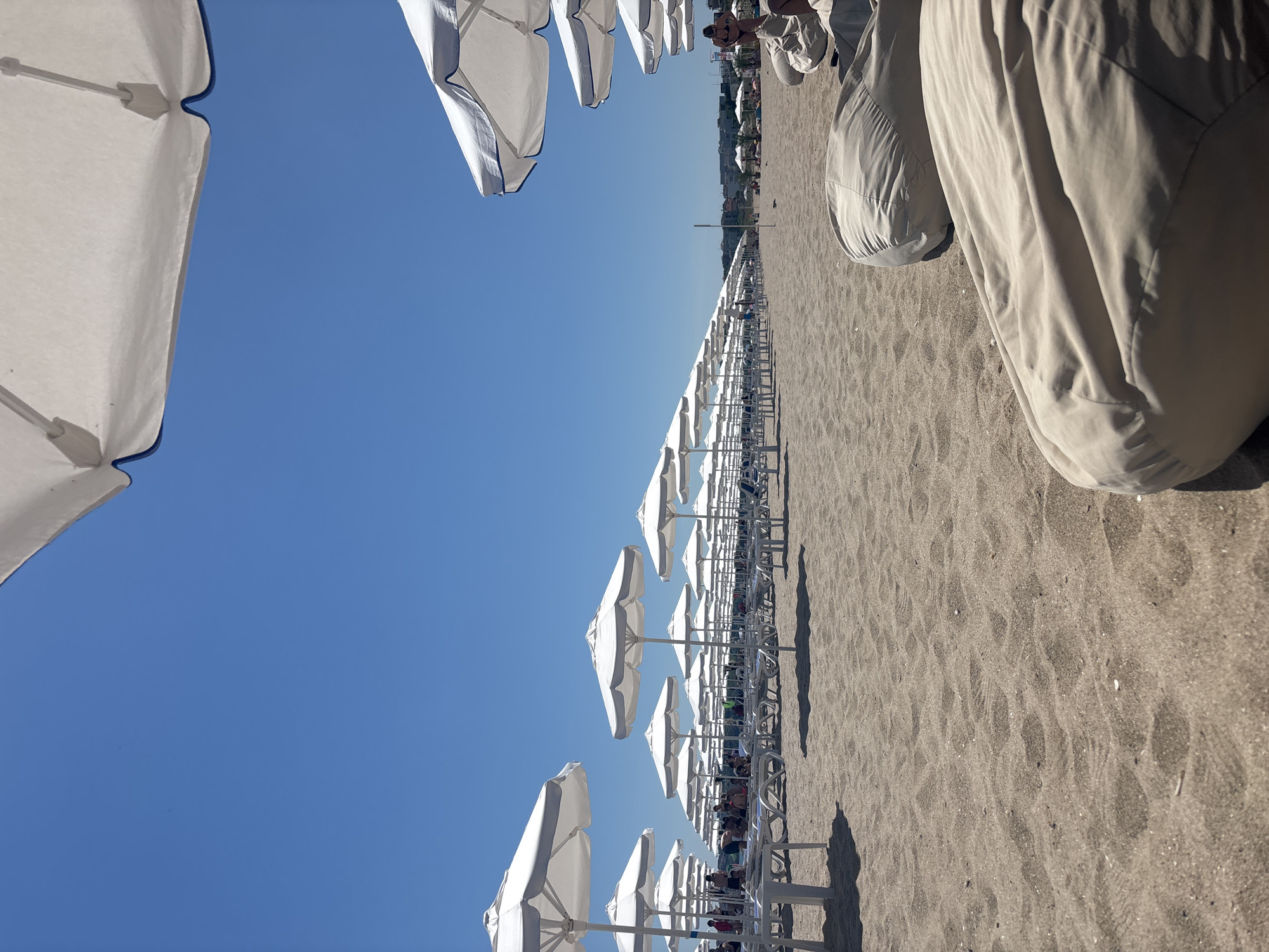 The image depicts a sun-drenched beach in Sozopol, Bulgaria. The scene is dominated by rows of empty white beach umbrellas and lounge chairs lined up on the sandy shore, suggesting a preparation for a busy day or the end of one. The bright blue sky indicates fair weather and a sunny time of day, likely midday.
In the foreground and to the left, a large white umbrella is partially visible, its fabric casting a shadow. Further down the beach, more umbrellas are arranged in neat rows, creating a strong sense of perspective that draws the eye towards the distant horizon. Some people are visible in the background, scattered amongst the loungers, their presence adding a human element to the otherwise orderly arrangement of beach furniture. The sand shows the textured patterns typical of a beach, with small flecks of shell or debris visible.
To the right of the frame, large, beige, beanbag-like chairs are positioned on the sand, also appearing empty. The overall impression is one of a popular tourist destination, ready to accommodate visitors for a day of relaxation by the sea. The lighting suggests it is a bright, clear day. No visible text is present in the image.