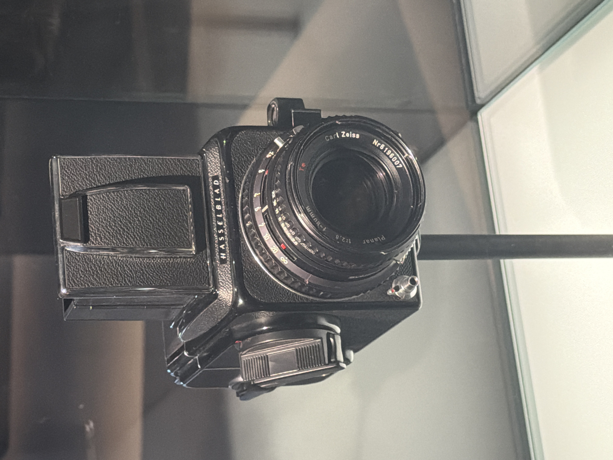 This is a close-up shot of a vintage Hasselblad camera, a high-end medium format camera known for its professional quality and durability. The camera is black with a textured leatherette grip on the film back. Attached to the body is a Carl Zeiss Planar 1:2.8 f=80mm lens. The lens has various rings for aperture and focus control, with markings visible. The serial number "Nr6196007" is clearly printed on the lens.
The camera appears to be displayed in a museum or exhibition setting, possibly behind glass, given the reflections on the surface of the lens and body. The background is out of focus, showing a muted color palette with hints of grey and white, suggesting an indoor, controlled environment. The lighting is diffuse, highlighting the details of the camera. There is no person visible in the frame. The scene is static, focusing solely on the camera as an object of display. The setting is identifiable as Rotterdam, Netherlands, though no specific landmarks are visible in the immediate vicinity of the camera. The overall impression is one of a well-preserved piece of photographic history.