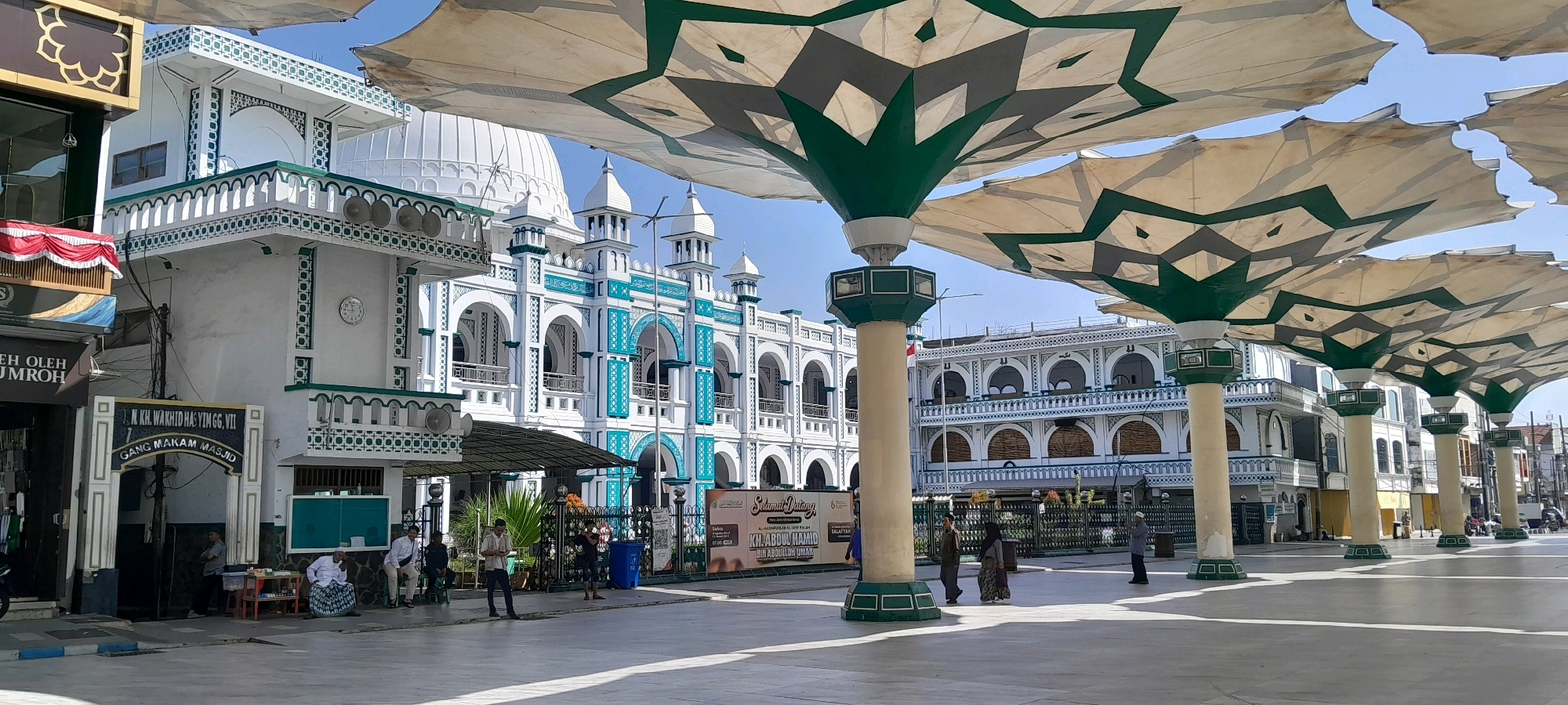 The image depicts the courtyard of the Masjid Agung Jami' Pasuruan in Pasuruan, Indonesia. Large, white and green star-patterned umbrellas on tall pillars provide shade in the foreground. Behind the umbrellas, a multi-story mosque building with white walls and green accents is visible, featuring arched windows and minarets. Several people are present in the courtyard, some walking and some sitting. One person stands near a sign that reads "GANG MAKAM MASJID". The overall scene is bright and sunny, suggesting it is daytime.