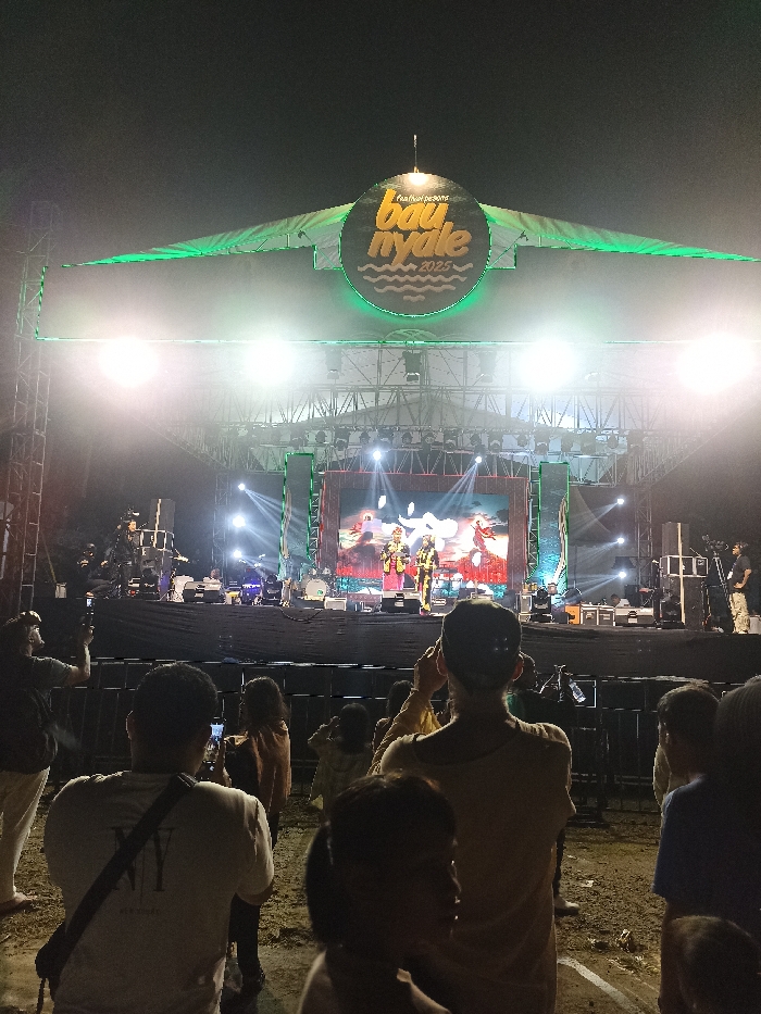 This image captures a vibrant night scene at the "Festival Pesona Bau Nyale 2025" in Kecamatan Pujut, Indonesia. A large, elevated stage serves as the focal point, brightly illuminated by numerous spotlights that cast beams through the night air, creating a hazy effect.
On the stage, several performers are visible, some dressed in what appear to be elaborate, possibly traditional, costumes. They are positioned in front of a wide LED screen displaying colorful graphics. Musicians and technical crew can also be seen on the stage and its periphery. Above the stage, a prominent circular sign features the festival's name: "festival pesona Bau Nyale 2025," with "Bau Nyale" written in large, distinct letters and a wavy line graphic underneath.
In the foreground, a crowd of people, including adults and children, is gathered, their backs mostly turned towards the viewer, indicating they are watching the performance. Many individuals are holding up smartphones, either recording videos or taking pictures of the event, demonstrating their engagement. The dark sky confirms the event is taking place at night. The overall atmosphere suggests a lively and captivating outdoor concert or cultural festival.