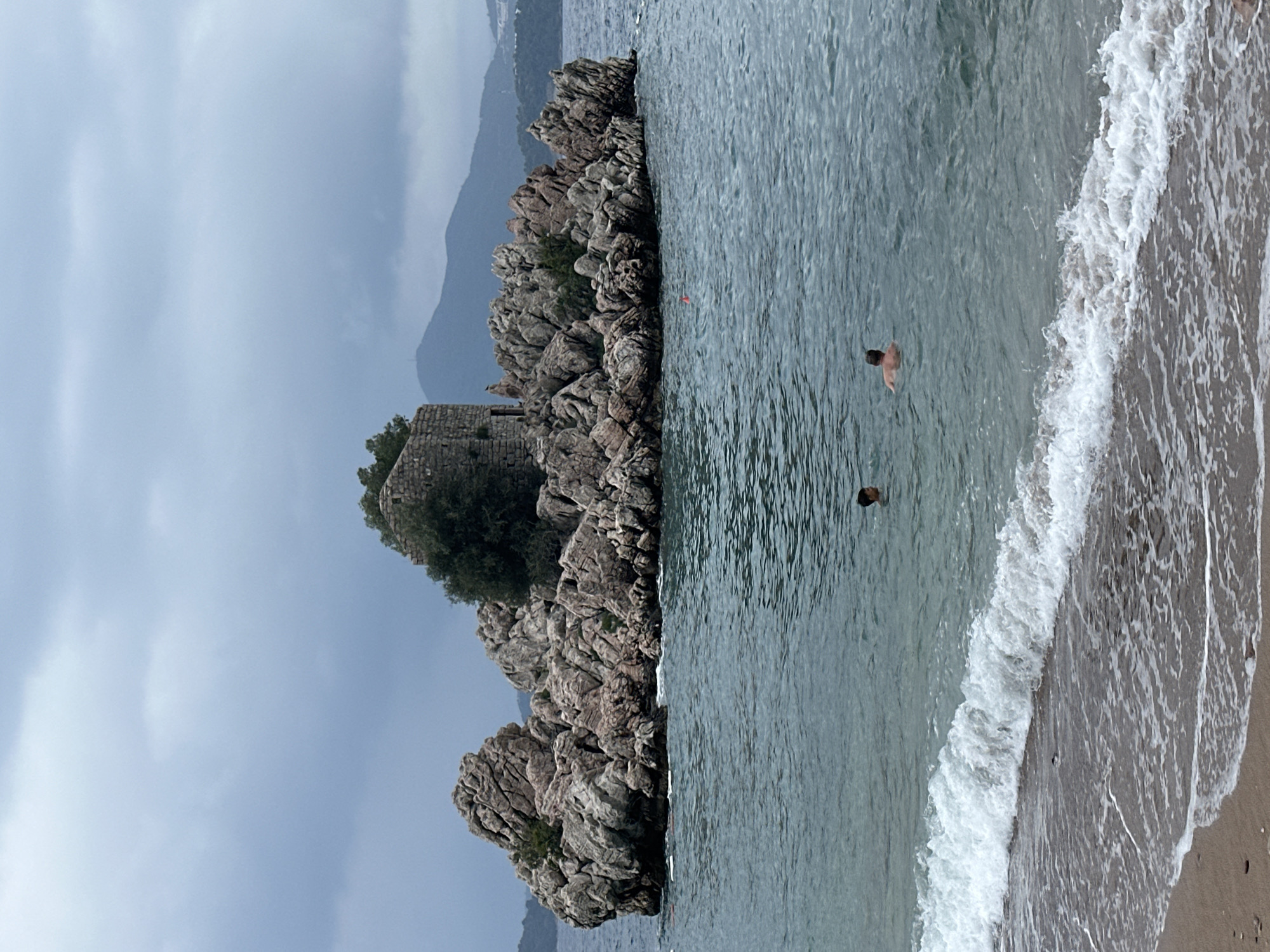 A serene coastal scene unfolds in Pržno, Montenegro, under an overcast sky. The image features a prominent rocky outcrop or small island on the left side, covered in rugged, greyish-brown rocks and sparse green vegetation, with trees at its peak. Perched atop this rocky formation, partially obscured by the foliage, are the weathered ruins of an old stone structure, possibly a tower or fort.
To the right of the rocky formation, the calm, greenish-blue sea extends, showing gentle ripples on its surface. Further to the right, a sandy beach meets the water, where soft, white-foamed waves are breaking along the shore. Two individuals are visible in the water, apparently swimming or wading. One person is closer to the beach, with their head and upper back visible, while another is further out, only their head showing above the waterline.
In the background, distant mountains rise under the diffused light of the cloudy sky. The overall impression is one of peaceful leisure on a calm, perhaps late afternoon or early evening, cloudy day. There is no visible text in the image.