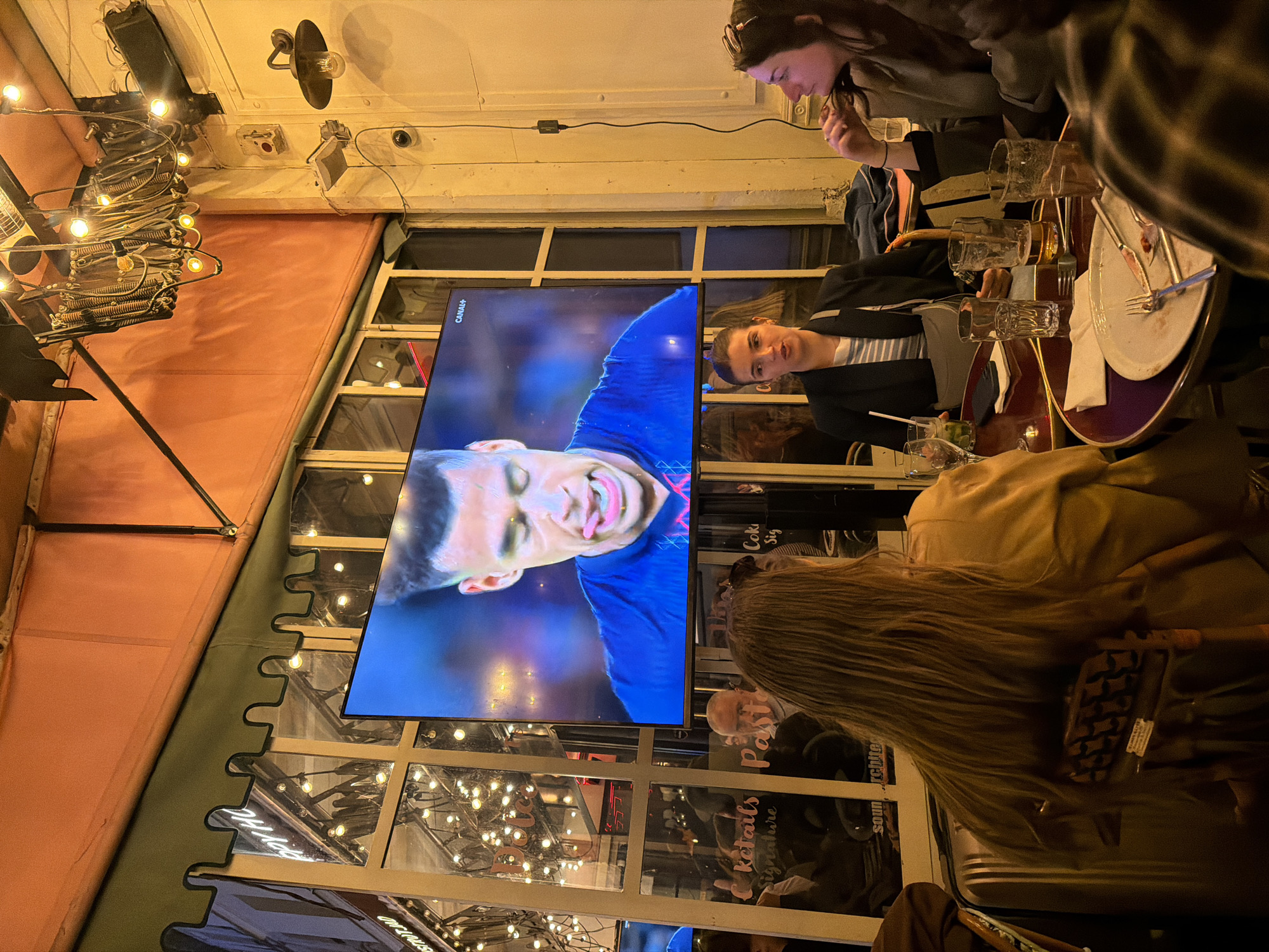 The image shows an interior scene, likely a restaurant or bar in Paris, France. A large screen displays a soccer game, with the player Kylian Mbappé visible. Several people are seated around a table. The table is cluttered with used glasses, plates, and cutlery. A woman in a tan coat has her back to the camera, another in a dark jacket is visible beside the TV. The surroundings are decorated with string lights, and the location is indicated by window signage with French words like "Cocktails signature" and "Restaurants Paris." The atmosphere suggests a casual, social setting, possibly during a sporting event broadcast.