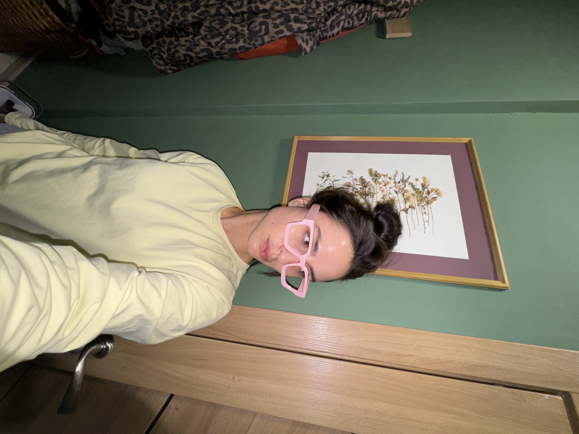 A woman with dark hair in a bun and pink glasses is positioned in front of a green wall with a framed floral picture. She is wearing a light yellow long-sleeved shirt. Part of a closet with an animal print garment is visible at the top left. Below the green wall is a light wood surface. A door handle is in the lower left. The location is indiscernible and there is no data to suggest it is in Krasnoyarsk, Russia.