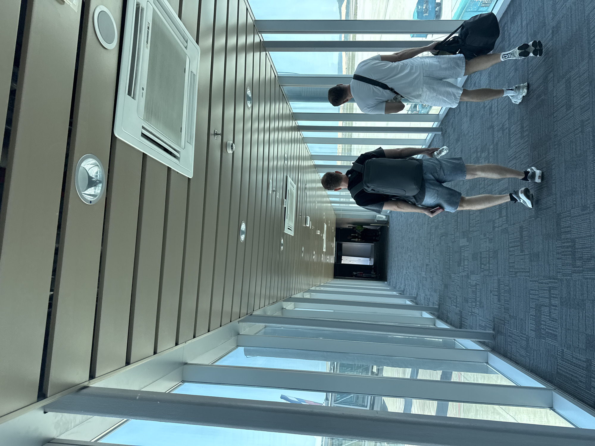 A passageway is viewed from an elevated angle, showing two men walking away from the viewer. The man closest to the camera is wearing a black t-shirt, grey shorts, and has a black backpack, holding a water bottle in one hand. Further away is another man dressed in white clothing with a black bag slung over his shoulder. The passage features a row of windows to the right of the frame. On the left, the wall is decorated with vertical panels, interspersed with circular lights and a rectangular air conditioning unit. The floor is carpeted in a grey patterned design. Outside the windows, the tarmac of an airport and parked aircraft can be seen.
