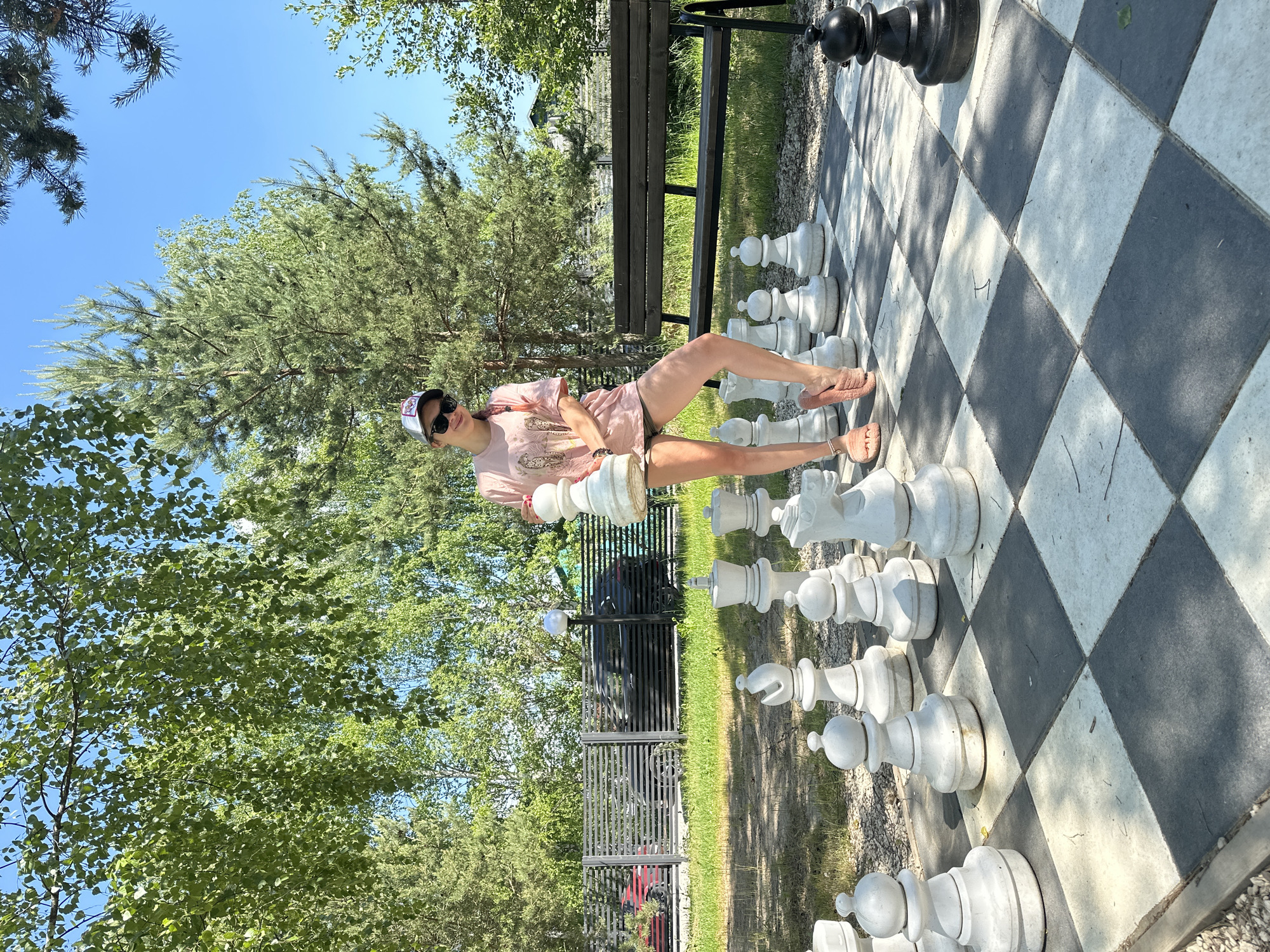 A young woman stands in the middle of a giant outdoor chessboard in Mikhalevo, Russia, holding a white chess piece, a queen. She is wearing a pink t-shirt, khaki shorts, sunglasses, and a baseball cap. The chessboard has large, white and black stone chess pieces. Behind her, a dark wooden bench is situated near a metal fence. Trees and a clear blue sky are visible in the background.