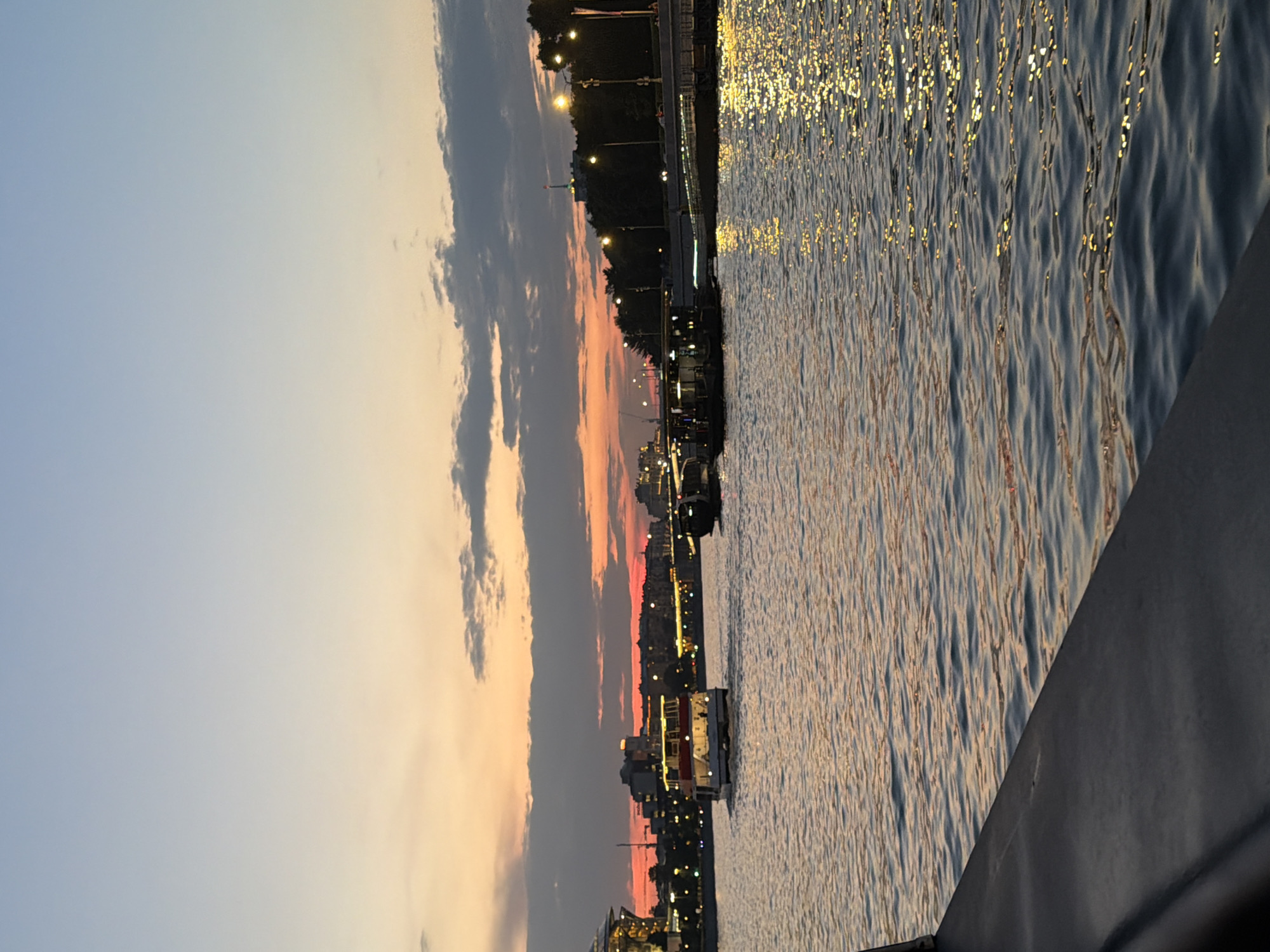 The image depicts a serene evening scene on the Moskva River in Moscow, Russia, likely during the golden hour or twilight. The sky transitions from a pale blue at the top to warm hues of orange and pink near the horizon, with scattered clouds. The river's surface is calm, reflecting the ambient light, with shimmering golden highlights from city lights and the sunset.
Several boats are visible on the water. In the mid-ground, a white passenger boat with red accents is moving from left to right. Further in the distance, other smaller boats and barges are also present. The shoreline is lined with trees and buildings, some of which are illuminated with warm lights, suggesting a developed urban area. Streetlights along the embankment are also visible, casting a warm glow. In the far left, a large, modern structure with glass facade and illuminated interior can be partially seen, possibly a building or part of a complex. The foreground shows a dark, solid structure, perhaps a railing or a part of a larger boat, from which the photograph is taken. There are no discernible people in the image.