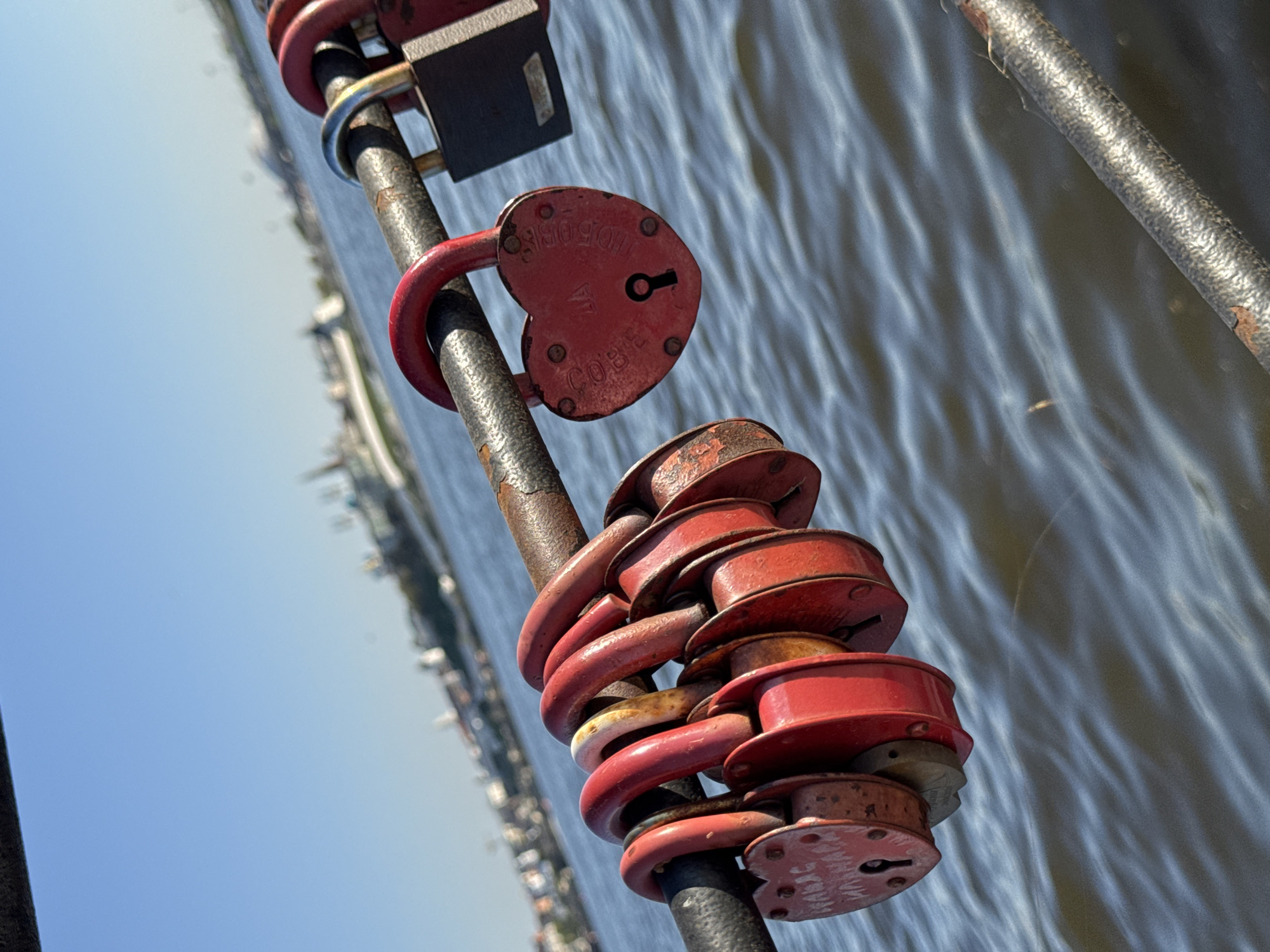 The image captures a cluster of red love locks attached to a metal railing. A single heart-shaped lock is also visible on the railing. In the background is the Kazanka river, and the blurred cityscape of Kazan, Russia. The scene is outdoors, with a view of the sky visible in the lower left portion of the frame.