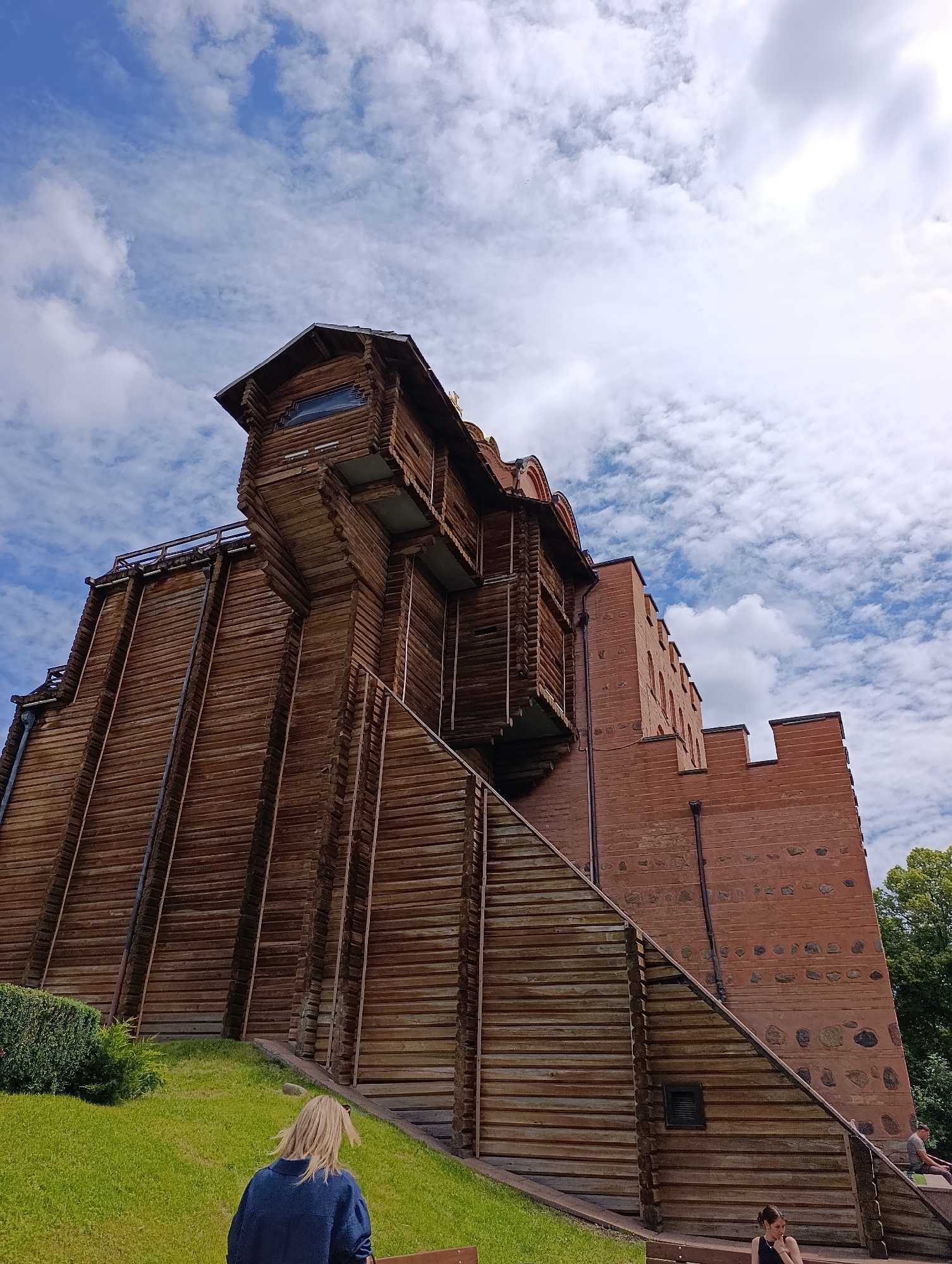 The image depicts the Golden Gate in Kyiv, Ukraine, a historical monument reconstructed in 1982. The structure is a prominent landmark, with its unique architectural blend of wooden fortifications and brickwork. The sky above is a vibrant blue with scattered white clouds, suggesting a pleasant day.
In the foreground, a person with blonde hair is seated, viewed from behind, wearing a dark blue jacket. To the right, another person is seated further down, also viewed from behind, wearing a black top. The area around the monument is well-maintained, with lush green grass and neatly trimmed bushes. The wooden facade of the gate is a dominant feature, with its intricate construction and weathered texture. The brick section of the gate showcases a more traditional fortress-like design. The perspective of the image is from a low angle, emphasizing the grandeur and height of the structure.