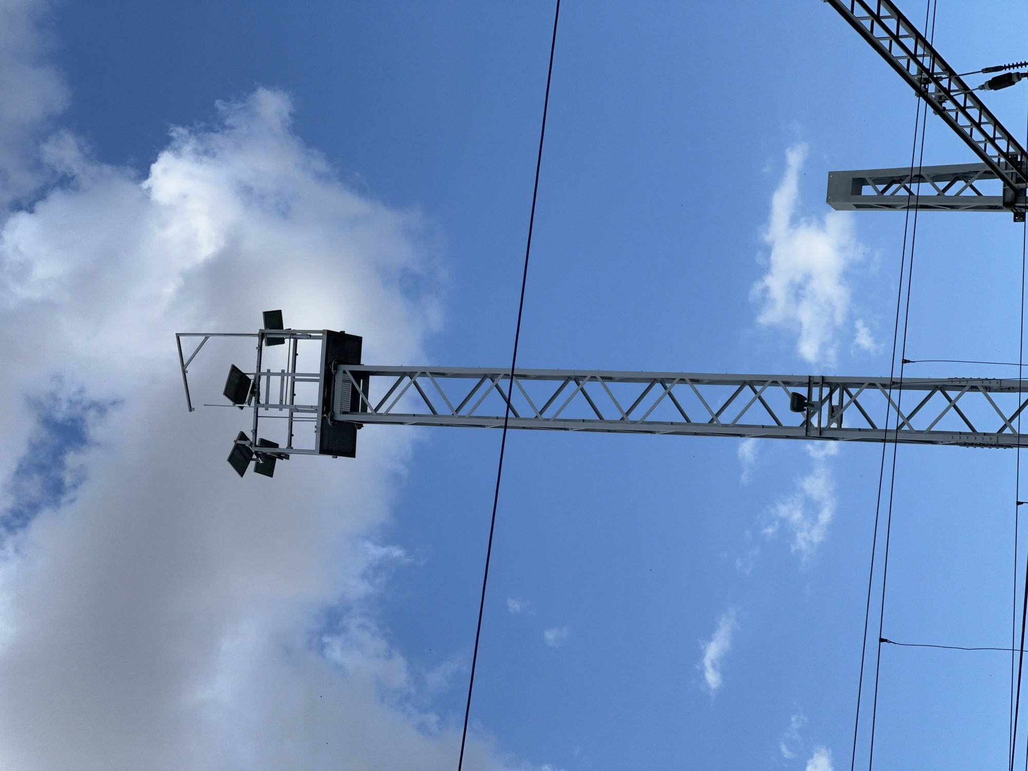 An outdoor, low-angle, full shot shows a tall, gray metal tower with lights on top. The tower is a lattice structure and is topped by a platform with several floodlights. The tower is situated in an open area with a bright blue sky and scattered white clouds. In the foreground, two dark, parallel wires run horizontally across the image, suggesting they are part of an electrical system. To the right of the main tower, another, shorter gray metal structure is partially visible. The overall scene appears to be part of a railway infrastructure, possibly in Újezd u Brna, Czechia. There are no people or animals visible in the image.