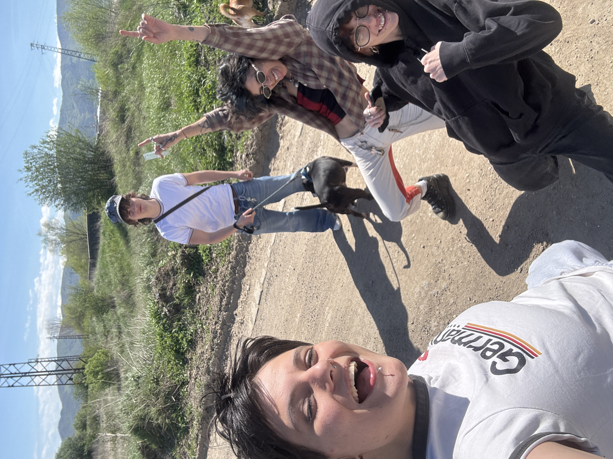A group of four young adults pose for a selfie on a dirt path in Zhivkovo, Bulgaria. The landscape includes grassy areas, a dirt path, and distant mountains beneath a blue sky. The path has vegetation and some trees alongside it, along with some utility poles.
The person taking the picture is in the foreground wearing a white t-shirt. Behind them are three other people. One is wearing a white t-shirt and jeans, holding a dog on a leash. The second person is wearing a plaid shirt and white pants, making a pointing gesture with one hand raised, and the third person is wearing a black hoodie. There's another dog in the background of the image.