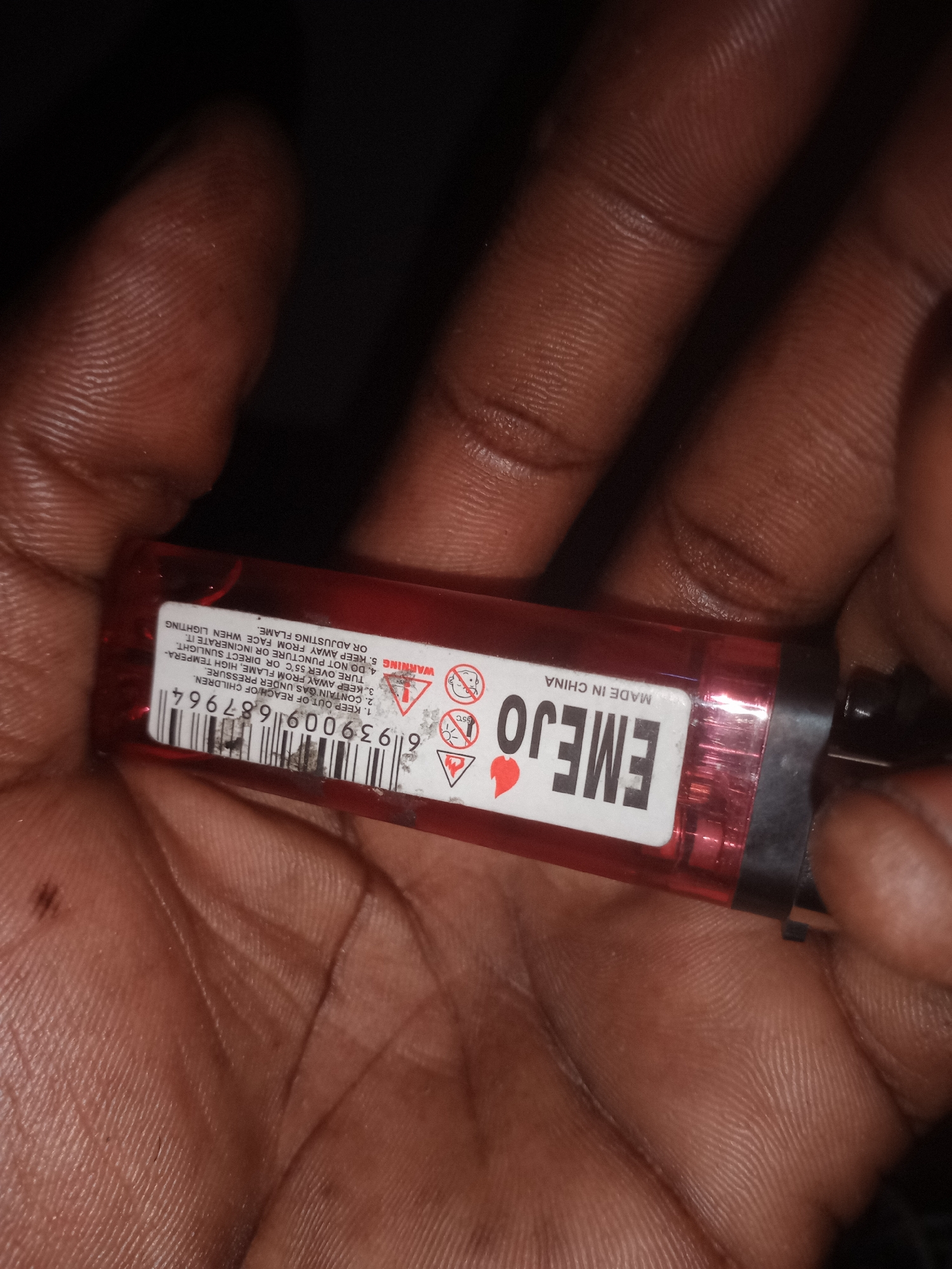 The image shows a close-up view of a person's hand holding a red disposable lighter. The lighter has white text and graphics on it. The text includes "EMEJO" prominently displayed, and further text reads "MADE IN CHINA." There is also a barcode and warning labels with symbols indicating flammability and keeping away from children. The background is dark and out of focus, suggesting an indoor or low-light environment. The skin texture of the hand, with visible fingerprints and lines, is detailed. The image is cropped to focus on the lighter and the hand holding it. There is no visible indication of the location being the city of Jalingo, Nigeria.