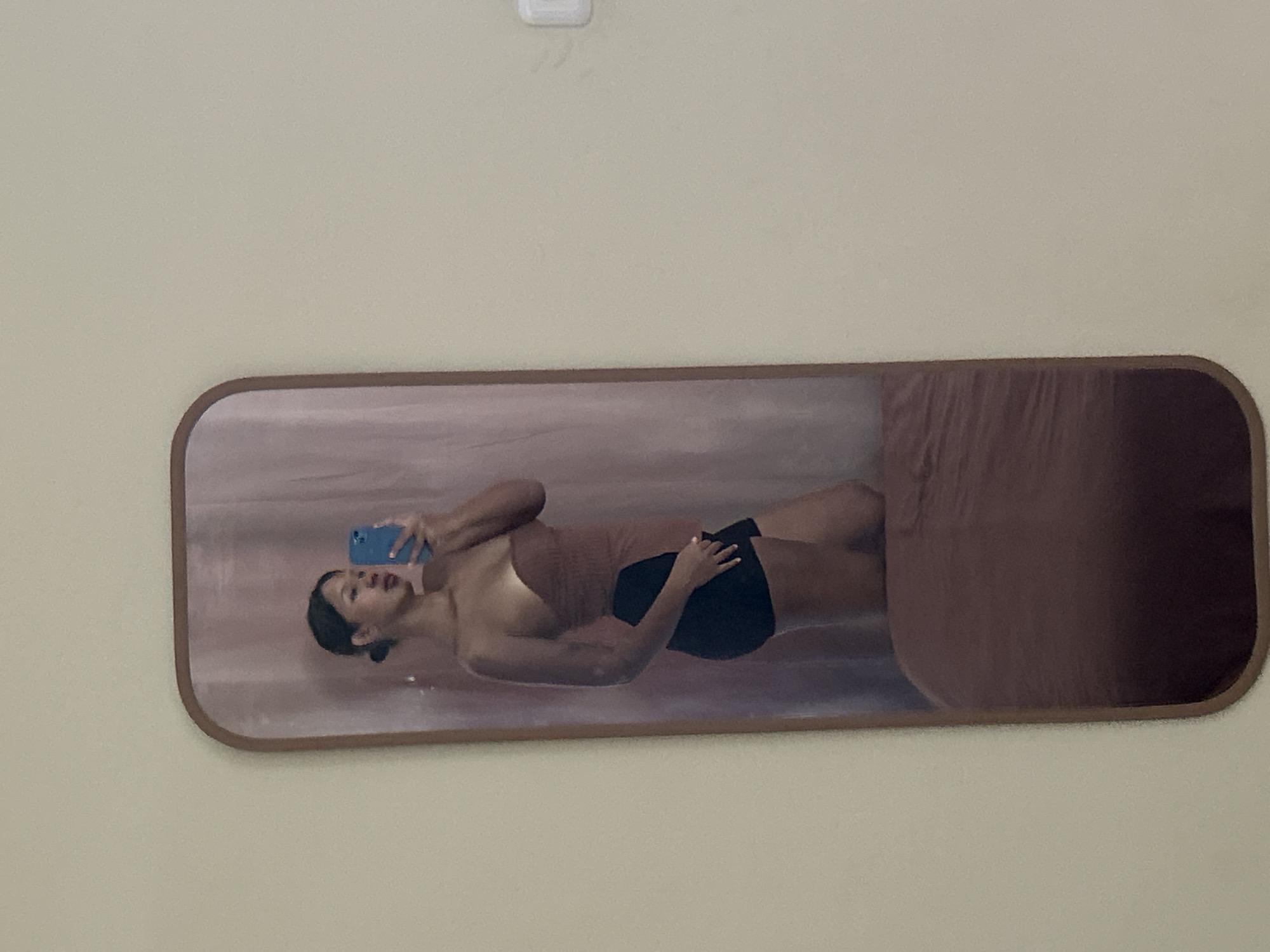 In a room with a light beige wall, a full-length mirror with a rounded top and a thin brown frame is positioned vertically. The mirror reflects a woman standing and holding a blue mobile phone in her right hand, as if taking a selfie. She is wearing a strapless, reddish-brown top and black shorts. Her hair is pulled back into a bun. Behind her, a pinkish-purple curtain is visible. The bottom part of the mirror reflects a bed covered with a reddish-brown comforter. The overall scene suggests a personal dressing area or bedroom. The specific location of Kuta, Indonesia, is not discernible from the image itself; this information would require external context.