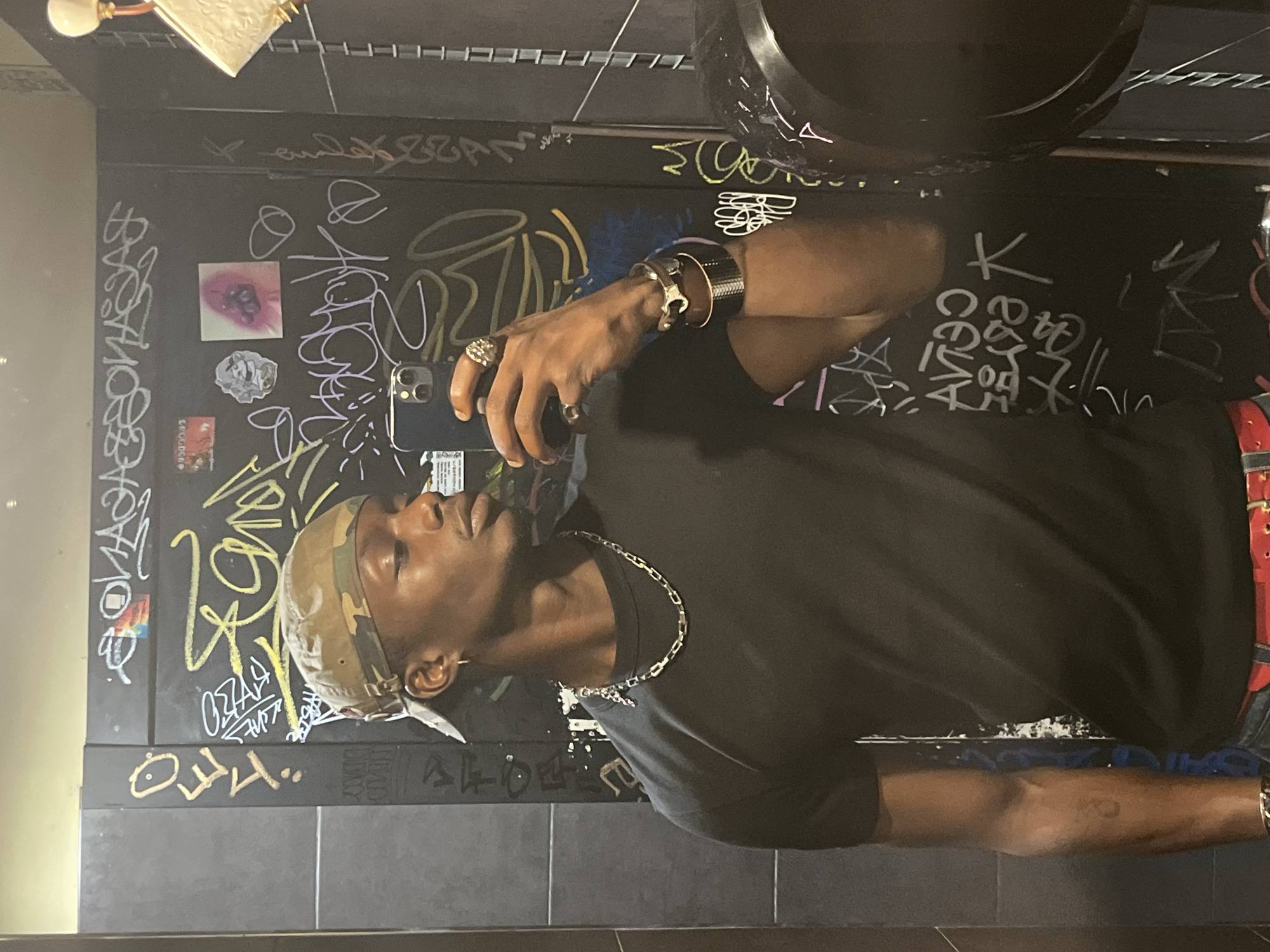 A young man wearing a black t-shirt, camouflage baseball cap, and red belt takes a mirror selfie in a restroom. The restroom walls are covered in graffiti. He is wearing a silver chain necklace, rings on his fingers, and a bracelet on his wrist. The image was taken at a Barcelona restroom.