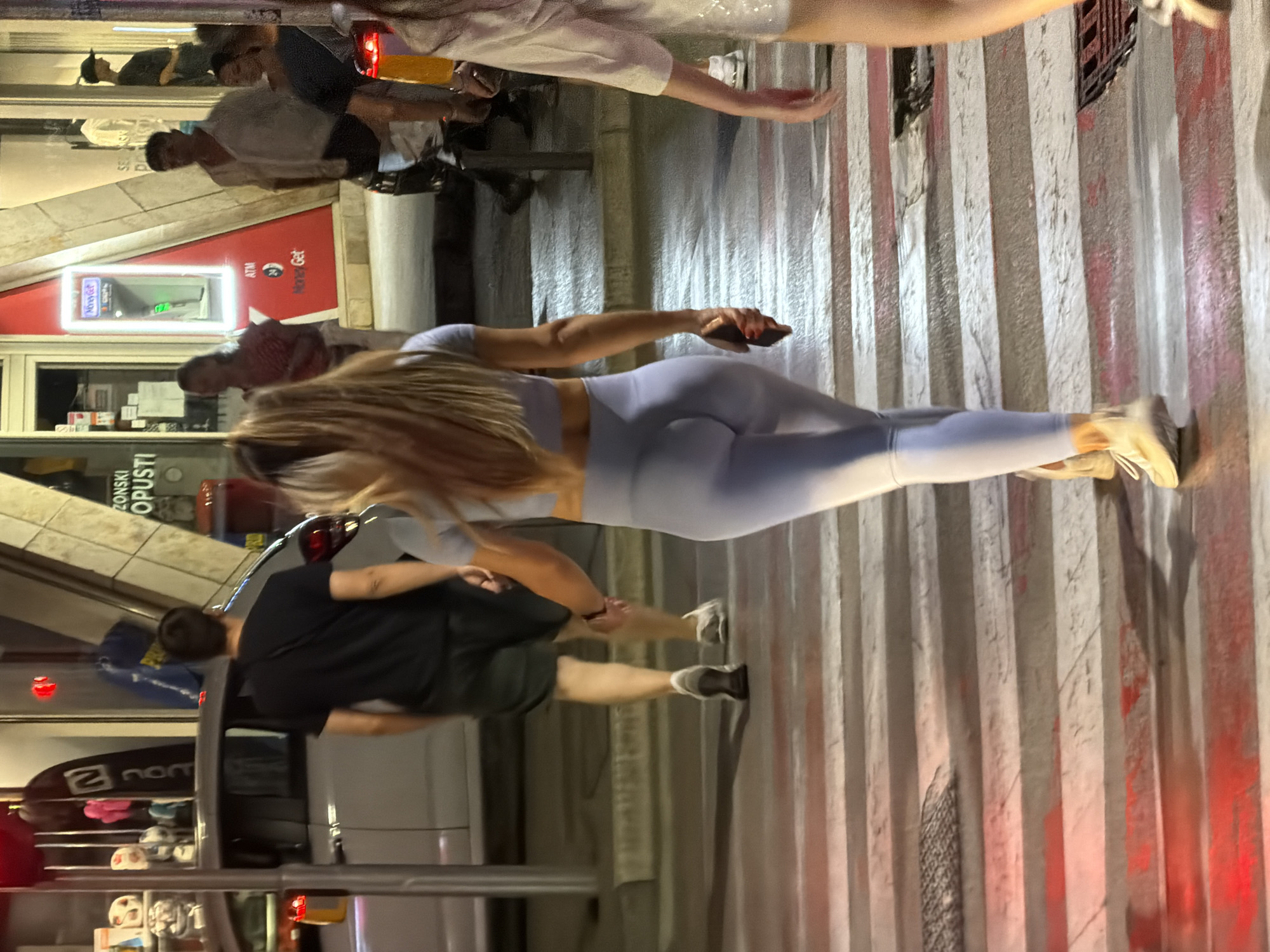 The image captures a nighttime scene on a pedestrian crosswalk in Budva, Montenegro. In the foreground, a woman with long blonde hair, wearing light blue athletic leggings and a matching top, is walking away from the camera across the crosswalk. She is holding a dark-colored mobile phone in her right hand. To her left, a man in a black t-shirt and dark shorts is also walking across the crosswalk in the same direction. Behind them, other individuals and a parked car are visible on the street. The background features storefronts, including one with a red facade and an ATM. The lighting suggests an urban environment with streetlights and illuminated signs.