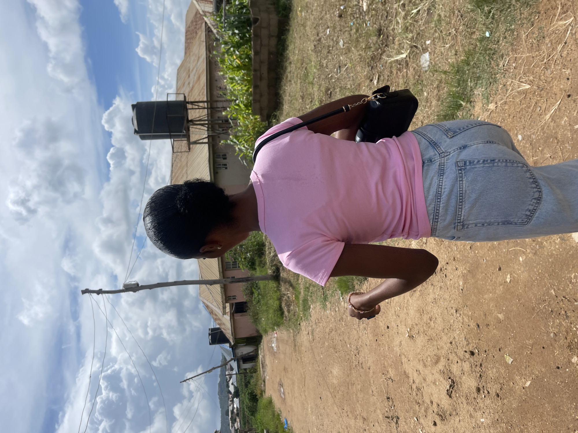 A young woman with dark skin and black hair pulled back walks away from the camera down a dirt road in Rafin Par, Nigeria. She is wearing a pink t-shirt and light blue denim jeans. She carries a black purse over her right shoulder. The road is lined with sparse vegetation and some trash. In the background, there are several buildings with corrugated metal roofs, a tall water tower, and power lines. The sky is blue with scattered white clouds. The overall impression is that of a rural or semi-rural setting.