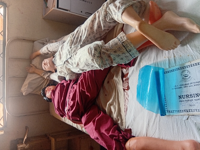 This image depicts a medical training simulation in a room at D.E.C. College of Nursing, located in Ochadamu, Ofu Local Government Area, Ejule, Kogi State, Nigeria.
Two full-body medical training mannequins are positioned supine on a single bed with a white metal frame. The mannequin on the left wears a maroon pajama set; its head rests on a pillow, and its lower legs and feet are exposed prosthetic components. The mannequin on the right wears a cream-colored, patterned pajama set; its head rests on a pillow, and both arms are extended upwards above its head. Its lower legs and feet are also exposed prosthetic components.
On the bed, between the mannequins' lower bodies, a blue plastic sheet partially covers a document titled "NURSING PROCEDURE MANUAL," which is further identified with the institution's name and a visible year "2022." The bed is covered with light-colored sheets. To the immediate left of the bed, a partial view of a wooden cabinet or shelf is visible. To the right of the bed, a light-colored wall features faint markings, and an overhead window with horizontal security bars allows ambient light into the room.