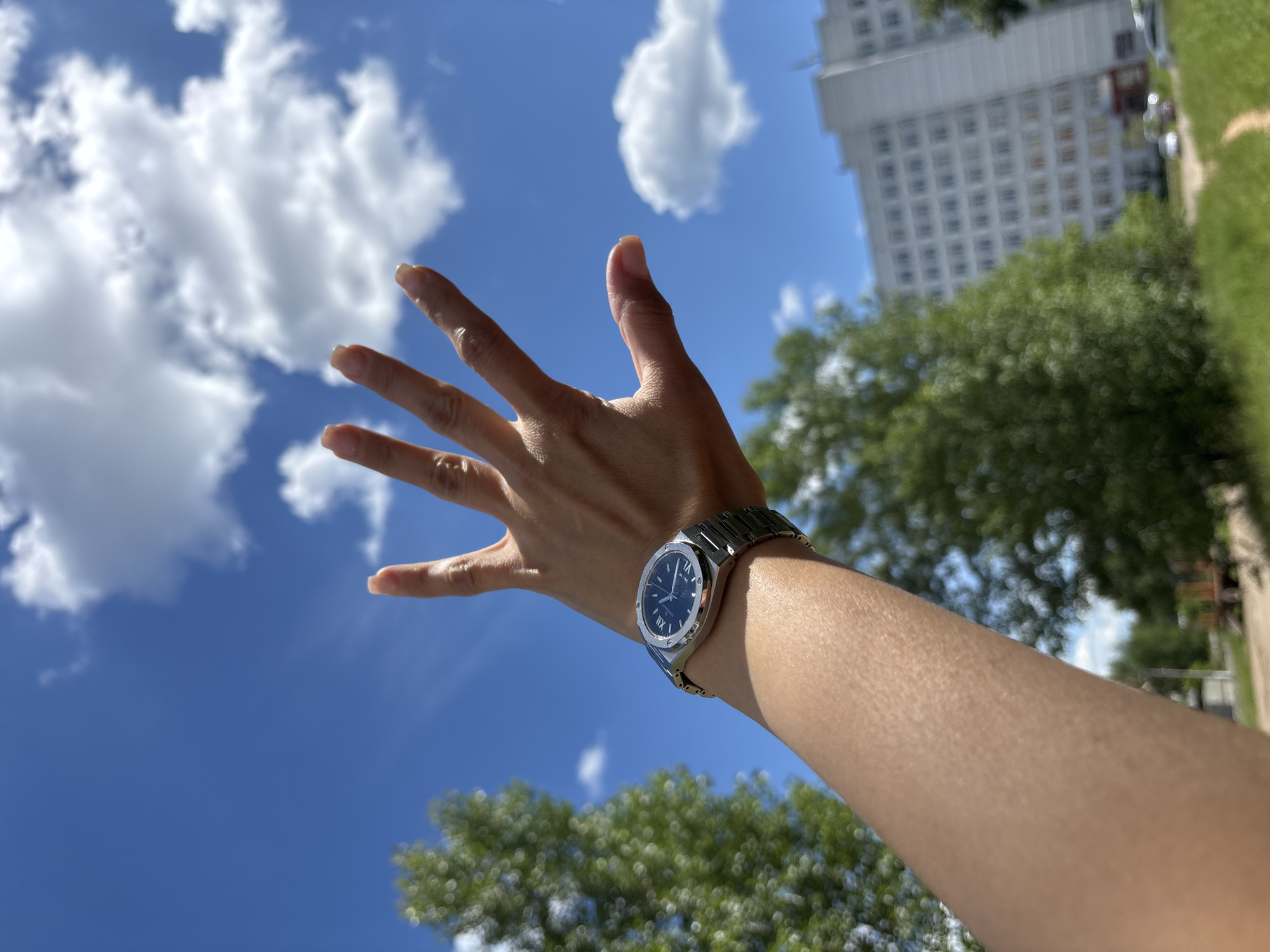 The image shows a person's arm and hand extended upwards towards a bright blue sky filled with fluffy white clouds. The hand is open, with fingers spread wide. A watch with a silver band and a dark face, featuring Roman numerals, is worn on the wrist. In the background, out-of-focus, are lush green trees, a modern multi-story building, and a grassy area. The scene suggests a sunny day, possibly during daylight hours, given the bright sky and shadows. The perspective is from below, looking up at the arm and sky. The overall mood appears to be one of reaching out or perhaps enjoying the outdoors. The location is identified as Chaikovsky, Russia.
