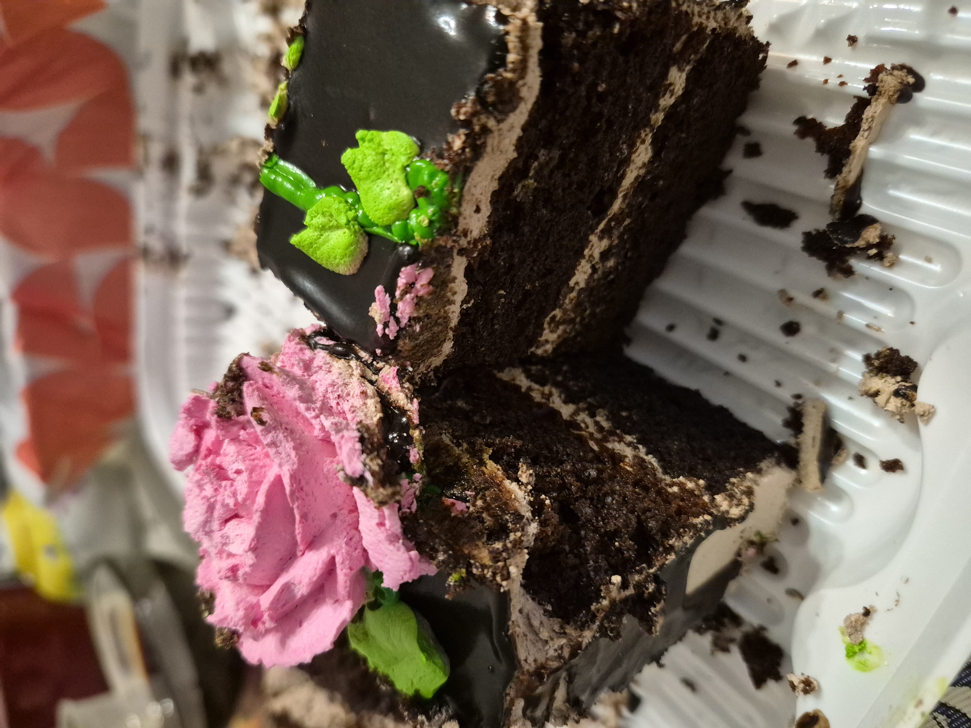 A close-up, slightly blurry, vertical shot shows two slices of chocolate cake on a white plastic tray. The cake on the left has a swirl of pink frosting that resembles a rose and a green frosting leaf. Crumbs and frosting are scattered on the tray. The cake on the right has a smooth dark chocolate glaze and two bright green frosting leaves. The background is out of focus, with hints of red and yellow objects. There is no indication of the city of Nyagan, Russia, or any people present in the image.