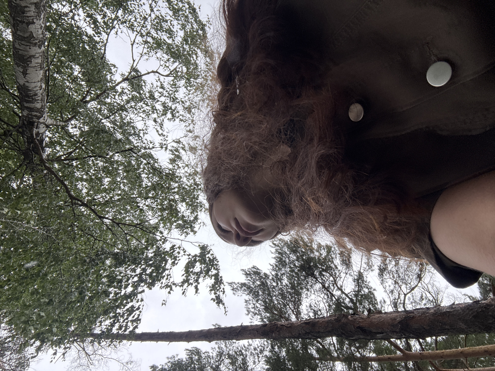 This is an extreme low-angle, upward-facing shot of a person lying down outdoors. The person's head is tilted back, and their face, with curly brown hair framing it, is partially visible. They are wearing a dark brown jacket with two visible silver snaps. The background consists of a dense canopy of green trees against an overcast sky, with some branches and the ground appearing in the foreground. The overall impression is one of being in a forest, possibly on the ground or on a low-lying structure. The lighting suggests it is daytime, but the cloudy sky indicates diffused light, possibly indicating overcast or rainy weather. The perspective makes it difficult to ascertain the exact activity, but the pose suggests relaxation or observation of the sky and trees. The image is from Chaikovsky, Russia.