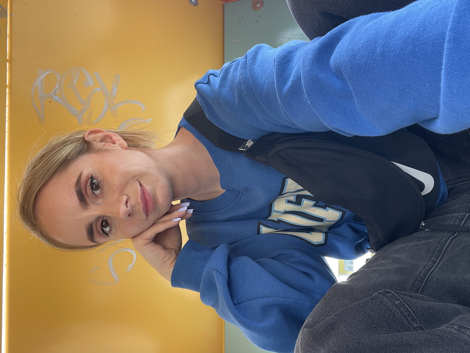 A blonde woman is sitting in front of a yellow surface covered in white graffiti. She is wearing a blue sweatshirt with white lettering, a black Nike fanny pack, and gray jeans. The location is unknown and cannot be determined from the image.