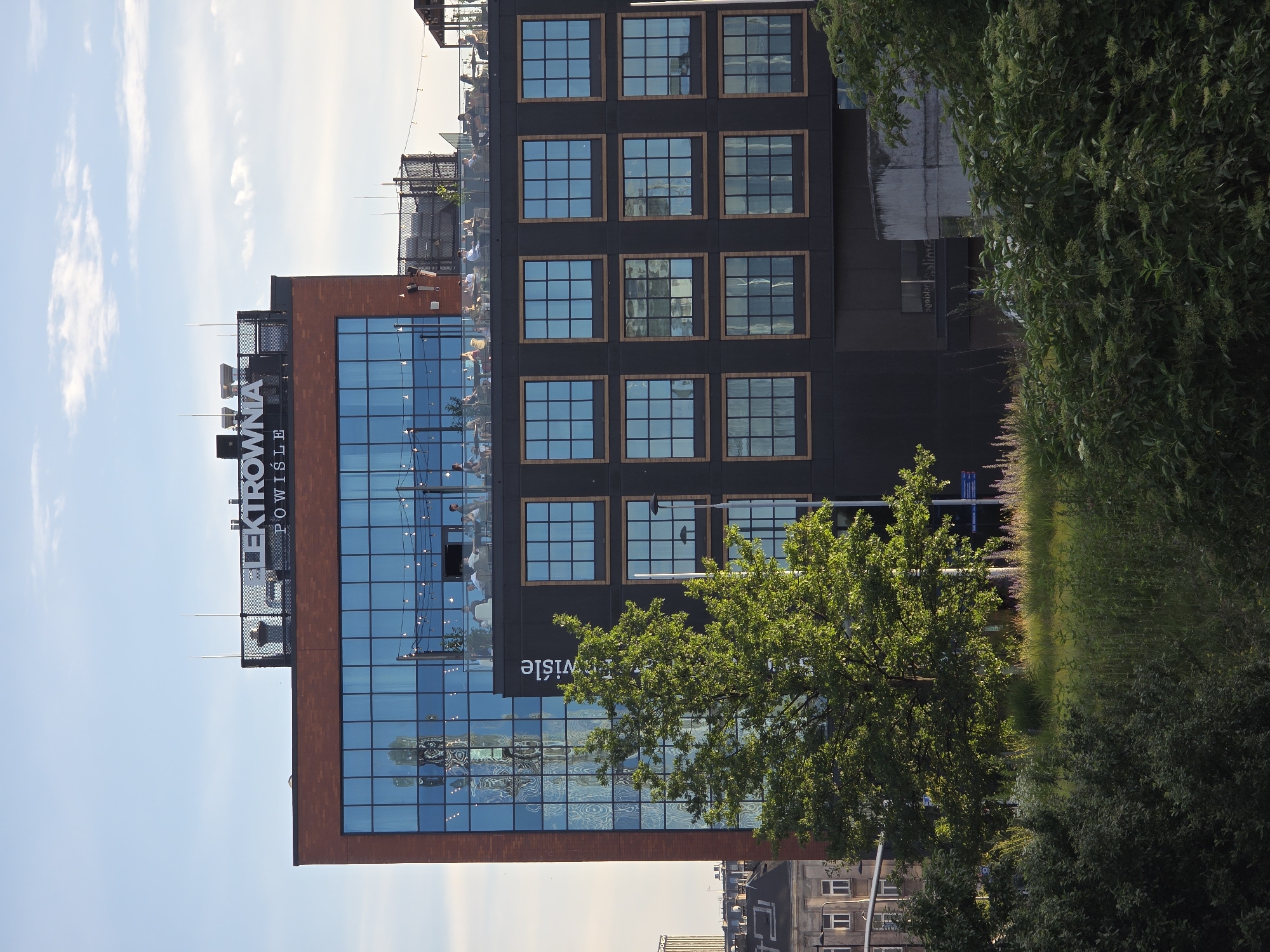The image depicts the ELEKTROWNIA POWIŚLE building in Warsaw, Poland, a multi-story structure with a facade of red brick and dark gray panels. The upper portion of the building features large, blue-tinted windows, reflecting a cityscape and the sky. A sign with "ELEKTROWNIA POWIŚLE" is visible on the rooftop. Several people are seated at tables on an outdoor terrace on the upper level, appearing to be dining or socializing. A mature, leafy green tree stands prominently in the foreground, partially obscuring the lower levels of the building, which are characterized by a grid of smaller, square windows with wooden frames. Lush green vegetation and foliage fill the bottom portion of the frame, suggesting a park or landscaped area surrounding the building. The sky is partly cloudy, with hints of a blue sky. The scene is captured during daylight, likely late afternoon or early evening due to the warm lighting.