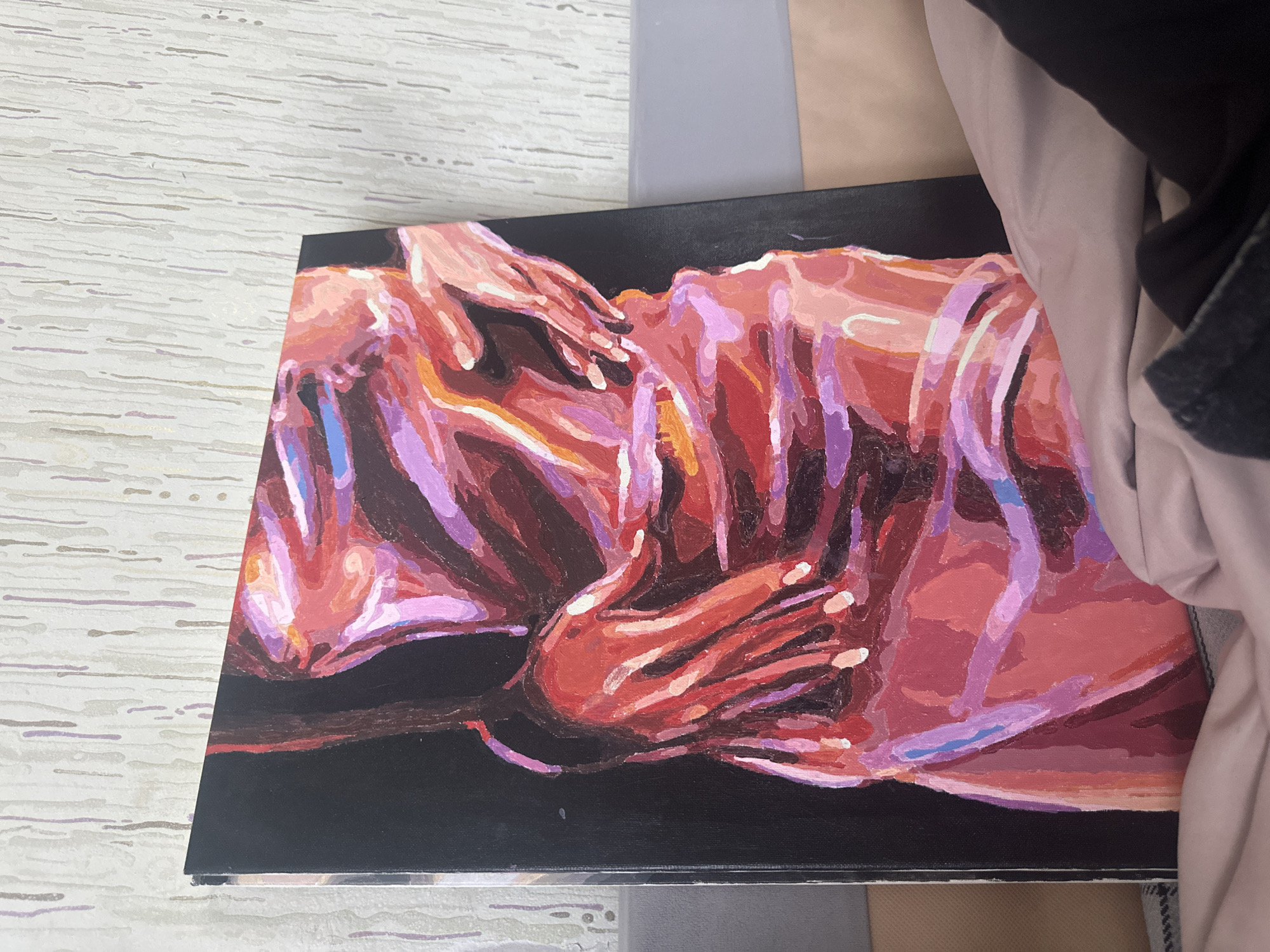 The image shows an acrylic painting on a black canvas. The painting depicts two figures intertwined, with a focus on hands and bodies. The color palette is dominated by shades of red, pink, and purple, with some orange and blue accents. The figures appear to be embracing. The painting is sitting on a surface that includes a light gray, textured pattern and a darker gray, plain area. A piece of fabric, possibly clothing, is visible in the background on the right side of the image. The location of Высокий, Russia is not determinable from the information presented in the image.