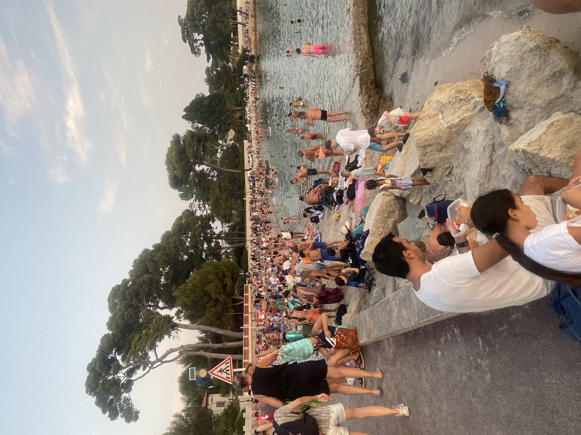 The image depicts a crowded beach scene in Antibes, France, likely during a summer evening. The photograph captures a wide view of the coastline, with a dense gathering of people enjoying the water and the beach. Pine trees and residential buildings line the background, indicating an urban coastal setting.
In the foreground, several individuals are visible, some sitting on rocks by the water's edge and others standing on a paved walkway. A man and two young girls are seated, with the man holding a container of food, seemingly engaged in a picnic. Further along the walkway, a group of people are standing, with one woman in a black tank top and shorts, and another woman to her left wearing a patterned top and white shorts.
The beach itself is populated by a large number of people, many of whom are in the water, swimming or wading. Others are sunbathing, sitting on towels, or walking along the shore. Beach chairs and various bags are scattered across the sand. In the water, several individuals are seen floating on inflatable devices.
The environment suggests a warm climate, with clear skies and calm water. The presence of palm trees and the architectural style of the buildings in the background are consistent with the Mediterranean coast of France. A pedestrian crossing sign is visible near the walkway, suggesting this area is accessible to the public and potentially near a road. The overall atmosphere is one of leisure and recreation, with a high density of beachgoers enjoying the seaside.